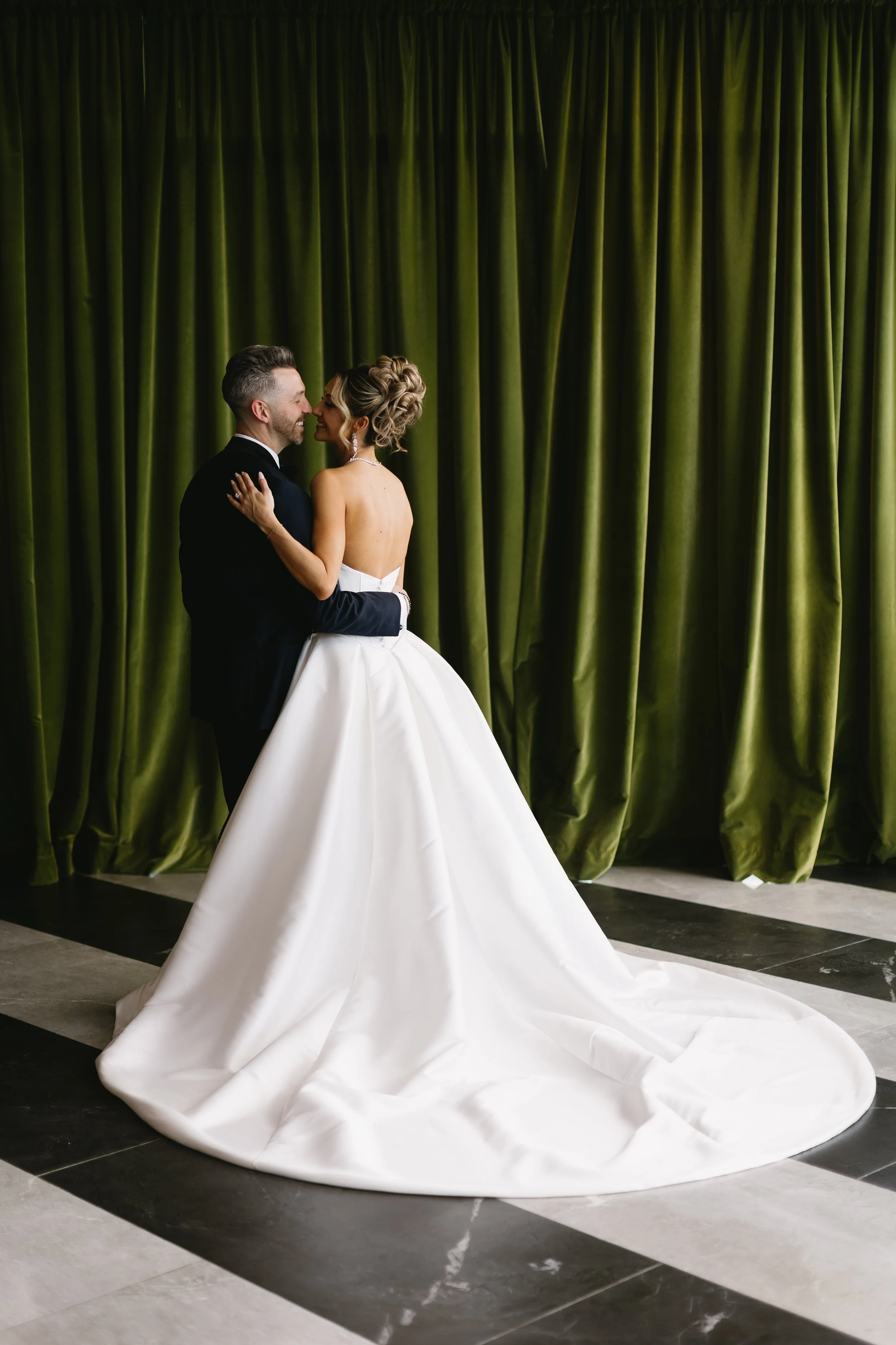 A bride and groom smiling and embracing in front of a green curtain, with the bride wearing a white wedding gown and the groom in a black tuxedo.