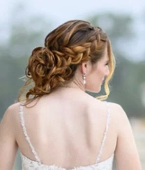 Side view of a woman with curly red hair styled in an elegant updo, wearing a white dress with thin straps, jewelry, and earrings, outdoors near a body of water.