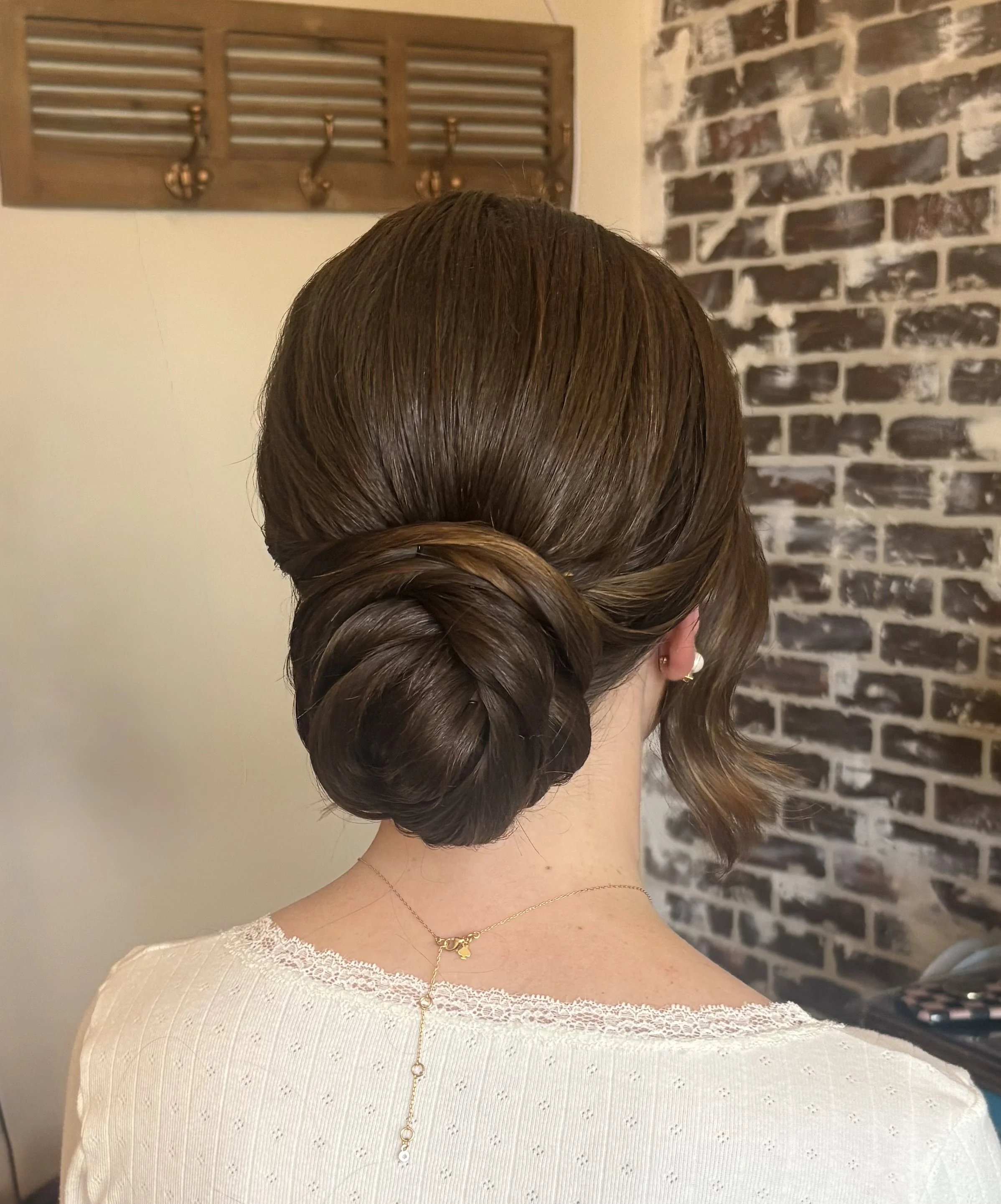 A woman with brown hair styled in an elegant twisted bun, dressed in a white lacy off-the-shoulder top, wearing a delicate gold necklace with a small pendant.