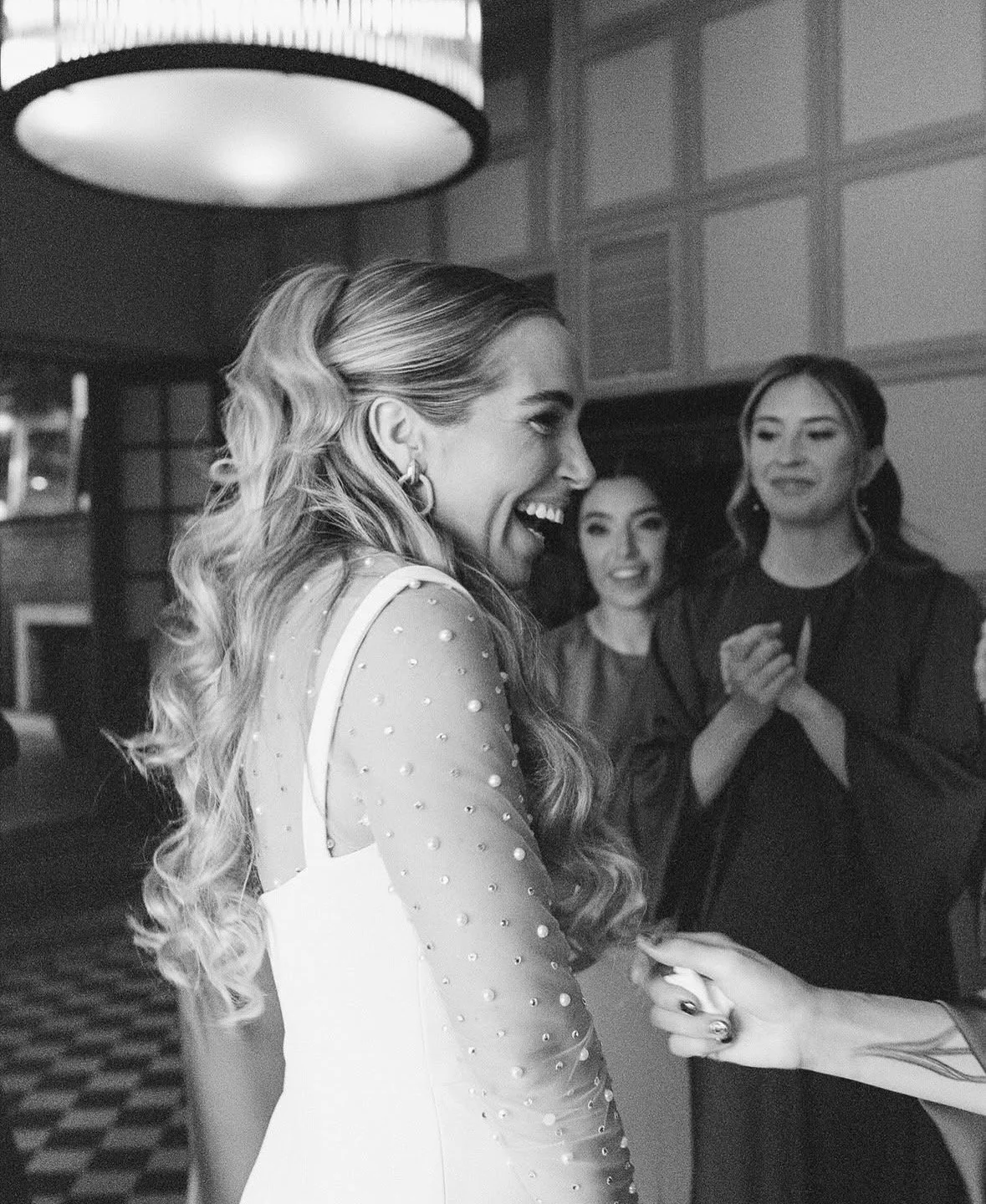 Woman with long, wavy hair smiling and holding hands with someone at a social gathering, with two other women smiling in the background.