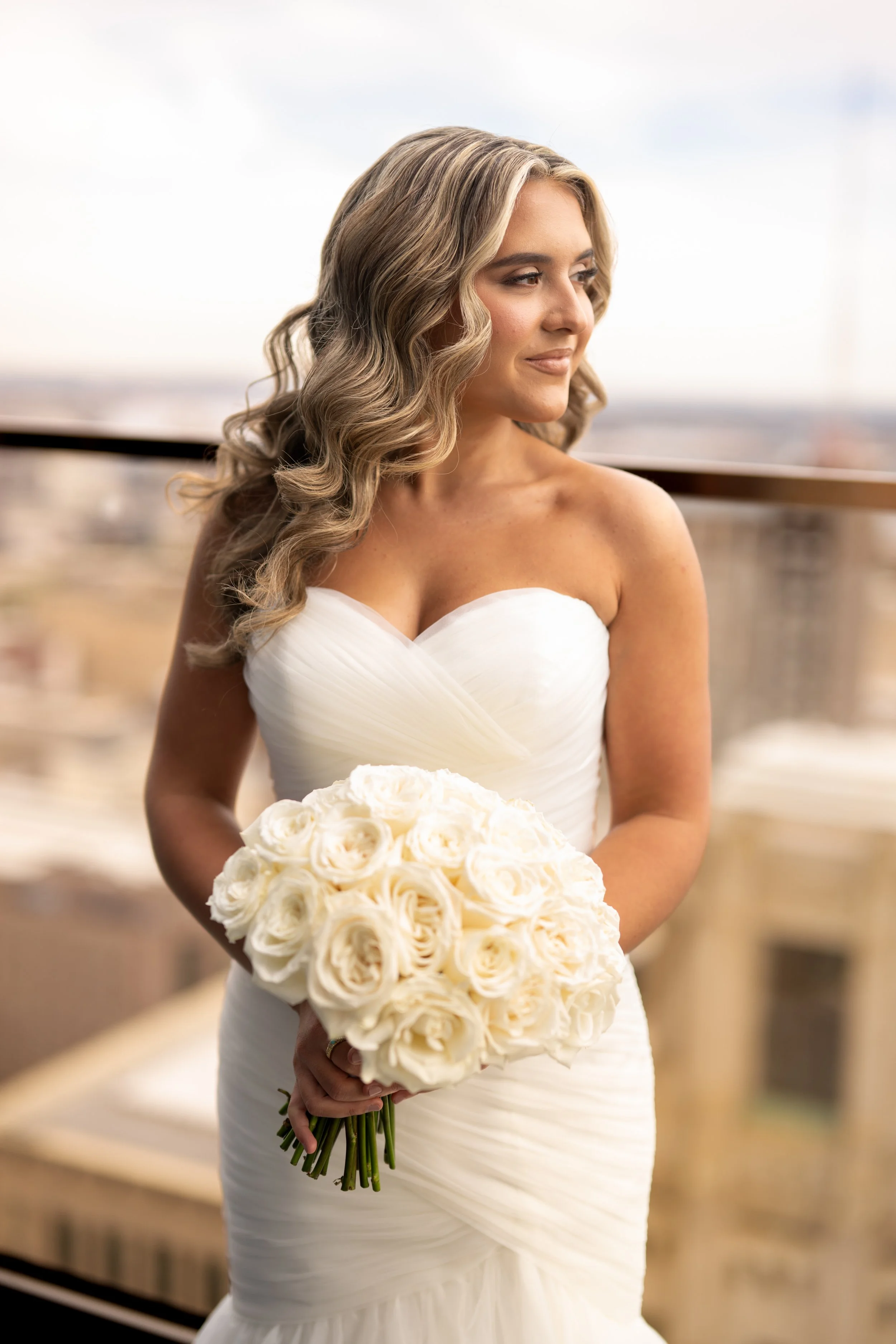 A bride with wavy blonde hair in a strapless white wedding gown holds a bouquet of white roses, standing indoors with a blurred background.