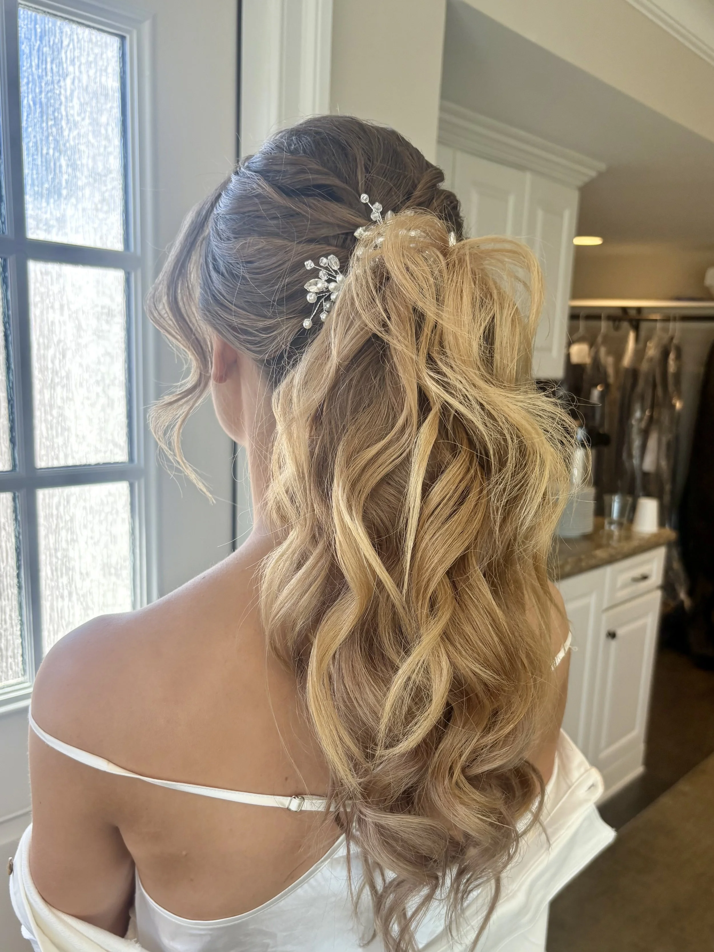 Side view of a woman with styled blonde and brown hair, adorned with a jeweled hair accessory, standing near a window with frosted glass, in a room with white cabinets and a closet in the background.