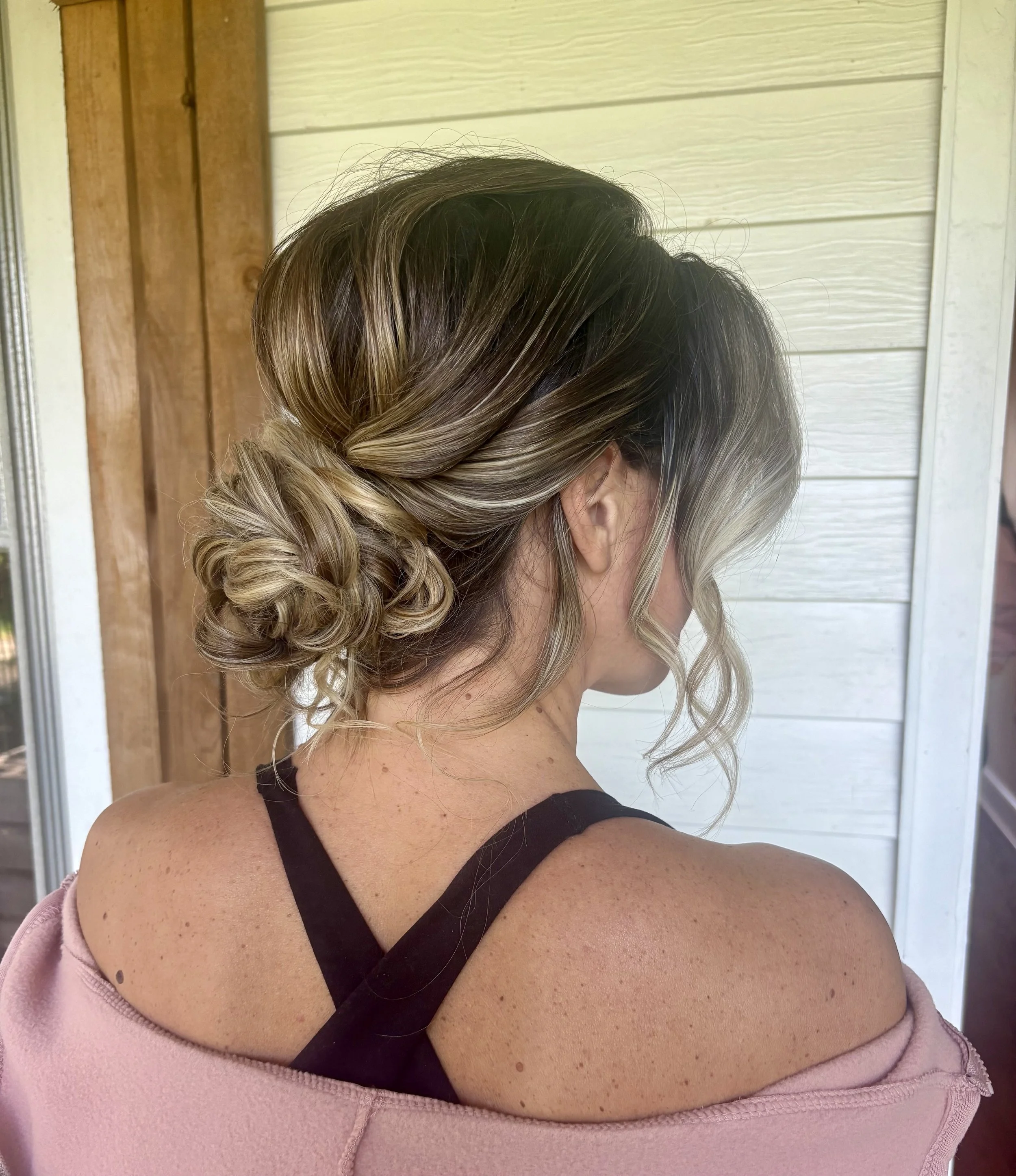 Close-up of a woman with styled blonde hair with darker roots, wearing a black bra and a pink jacket slipping off her shoulder, revealing her tan skin and freckles. The background features a wooden wall and siding.