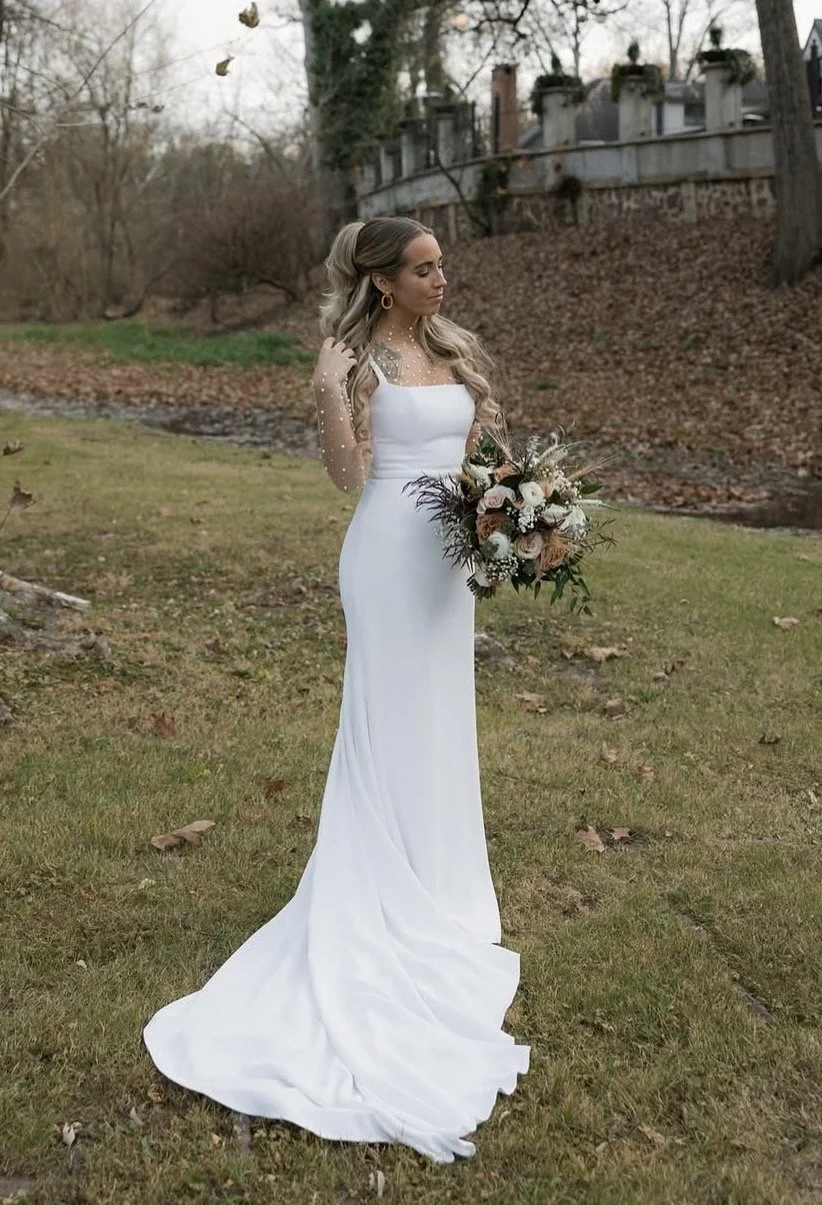 A woman in a white wedding dress holding a large bouquet of flowers outdoors in a park or garden with trees and a building in the background.