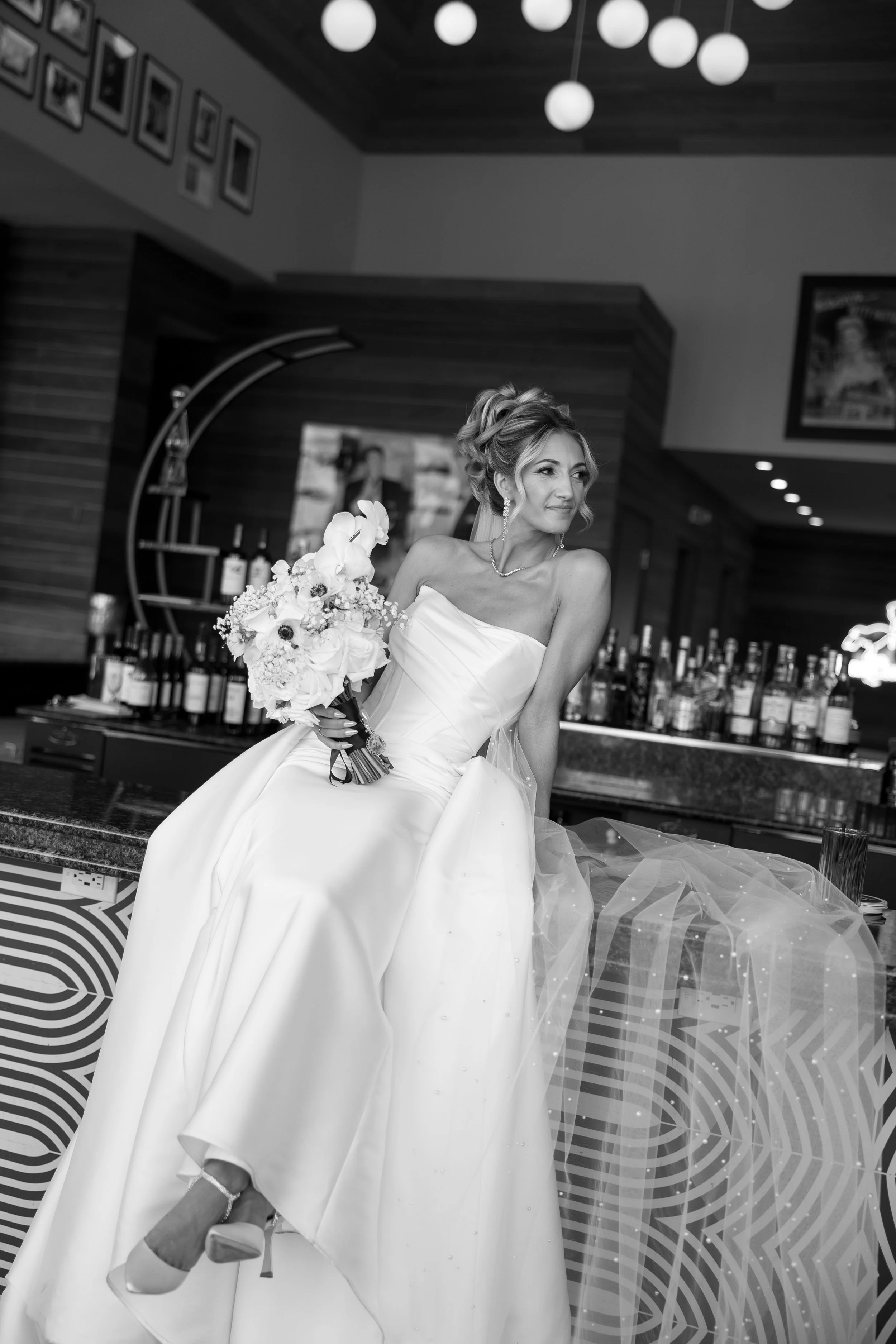 A bride in a strapless wedding gown holding a bouquet of flowers, sitting in a modern bar or lounge area with bottles of alcohol in the background.