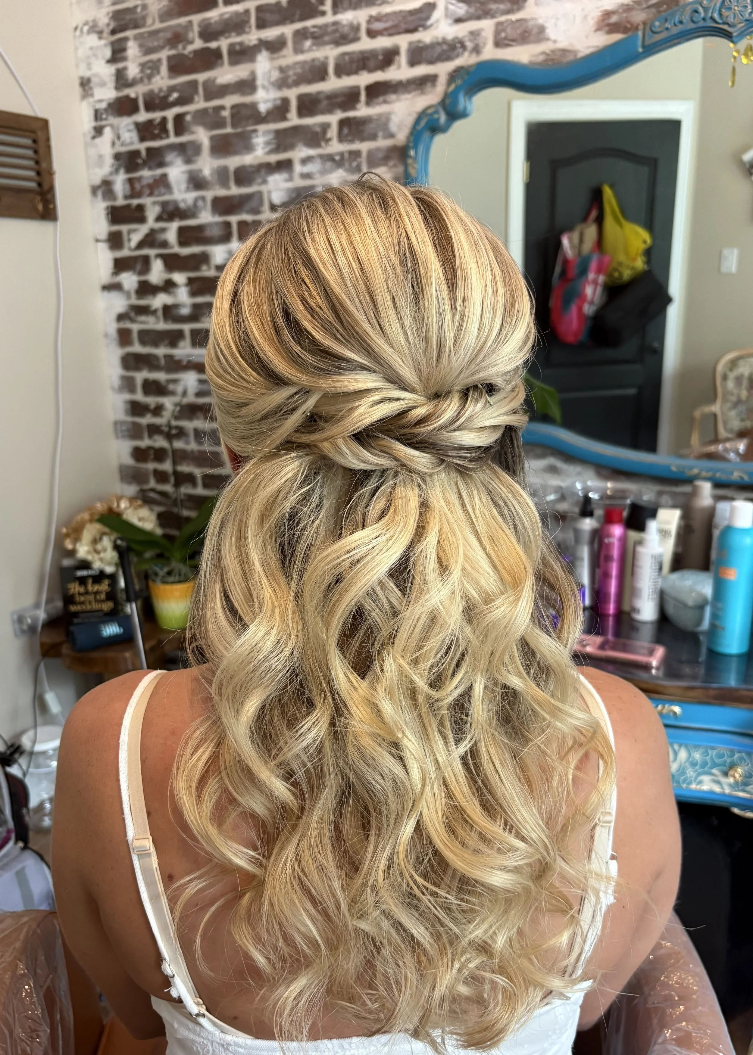 Back of woman's head showing long, blonde wavy hair styled in loose curls with a twisted braid section at the top, set against a brick wall and a mirror reflecting part of a room with bags and various items.