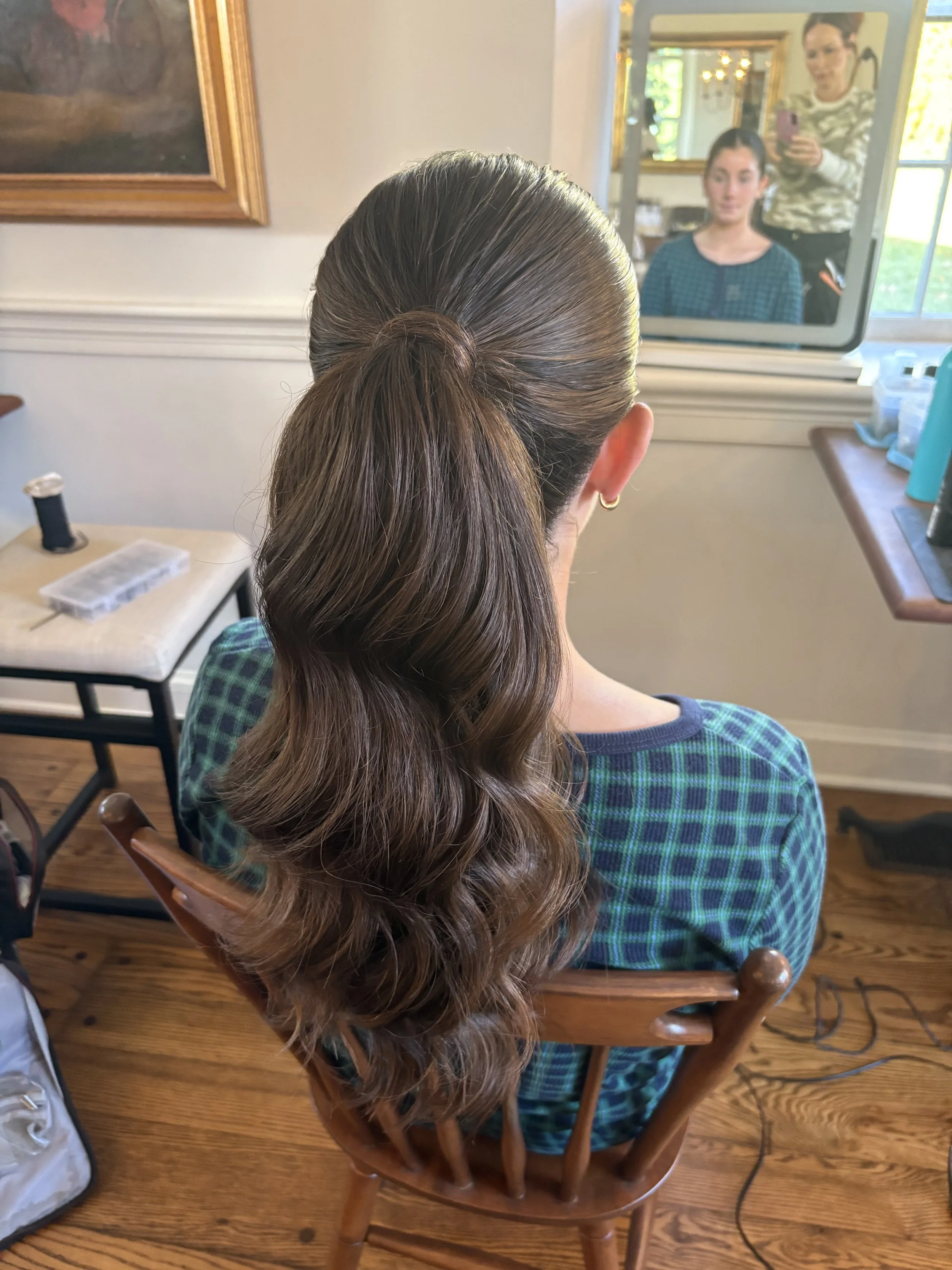 Woman with long brown hair styled in a loose ponytail is sitting on a wooden chair in front of a mirror, with a person taking a photograph in the background.