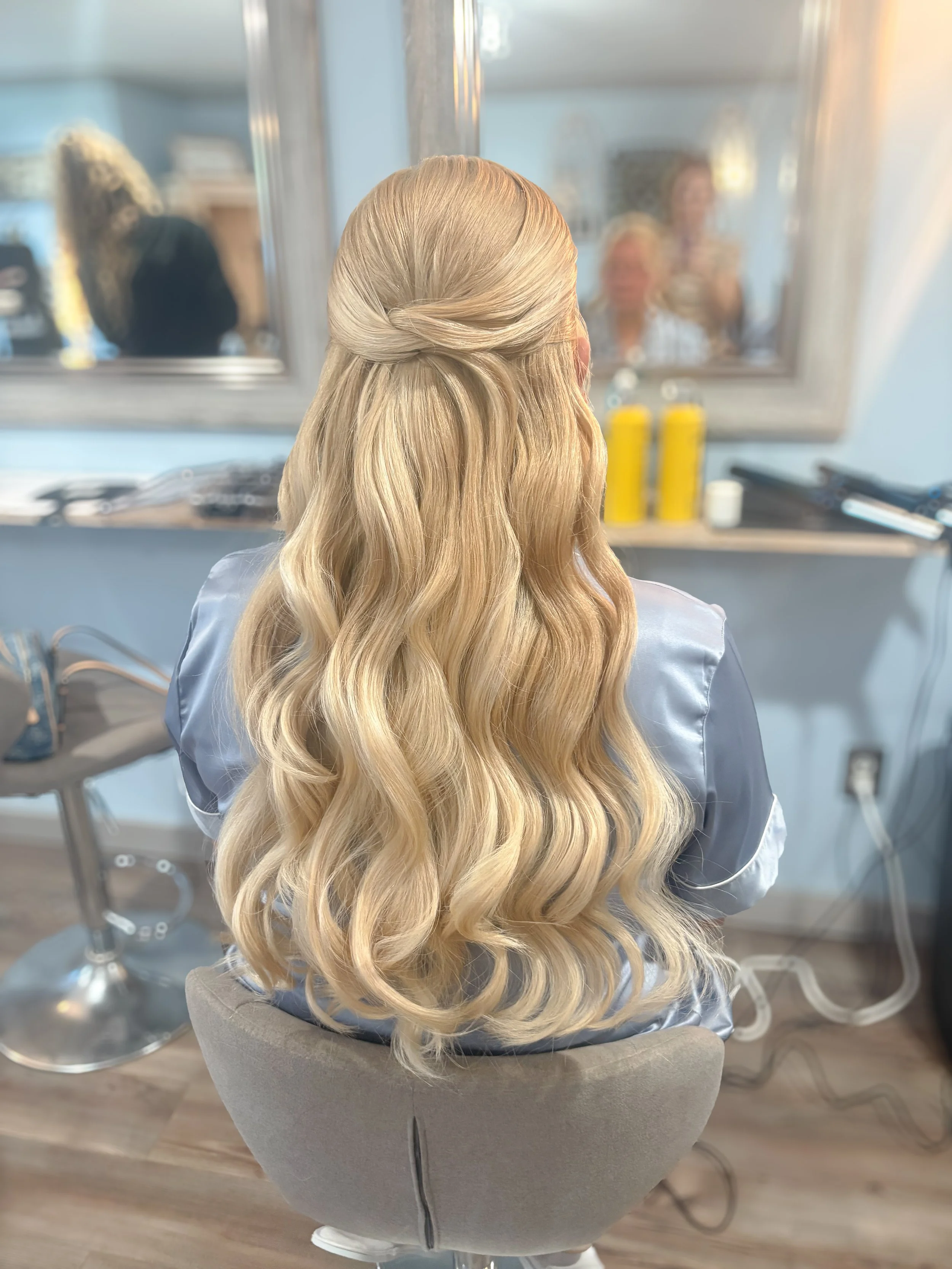 Back of a woman with long, blonde, wavy hair styled in loose curls, sitting in a salon chair in front of a mirror.