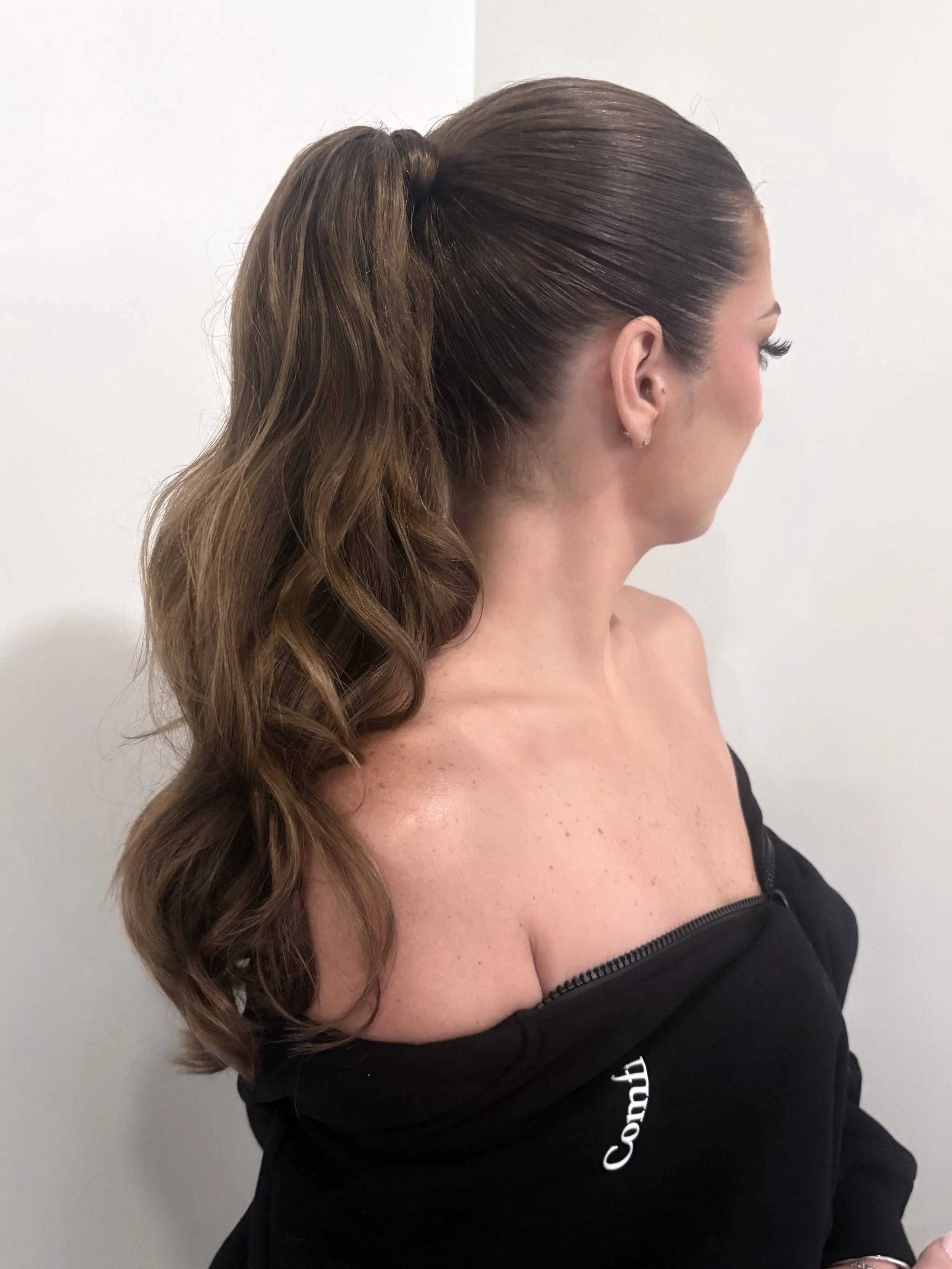 A woman with long, wavy brown hair styled in a high ponytail, wearing a black off-shoulder top and earrings, standing against a plain white wall.