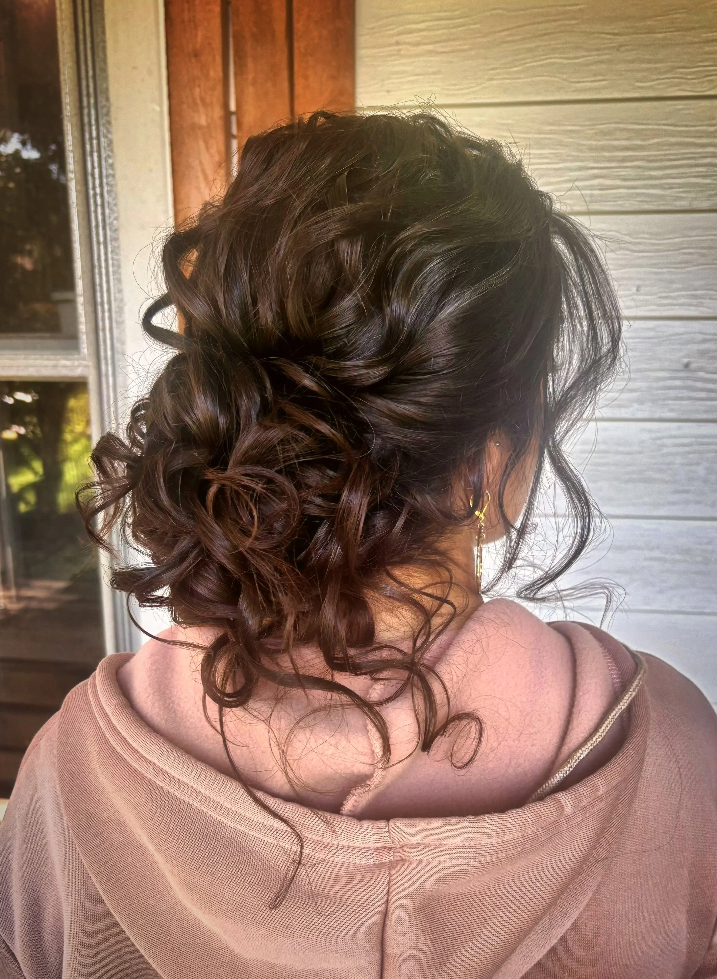 A woman with dark brown, curly hair styled in loose ringlets, wearing a beige hoodie, standing outdoors near a wooden wall and window.
