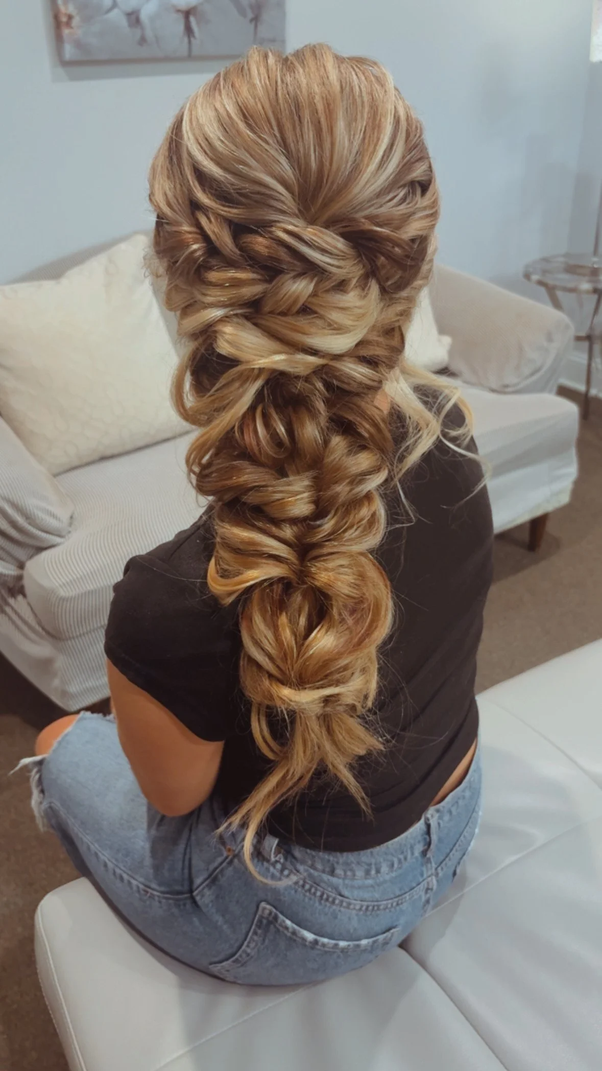 Back view of a woman with long, blonde, curled hair styled in an intricate braid, sitting on a white ottoman in a living room.