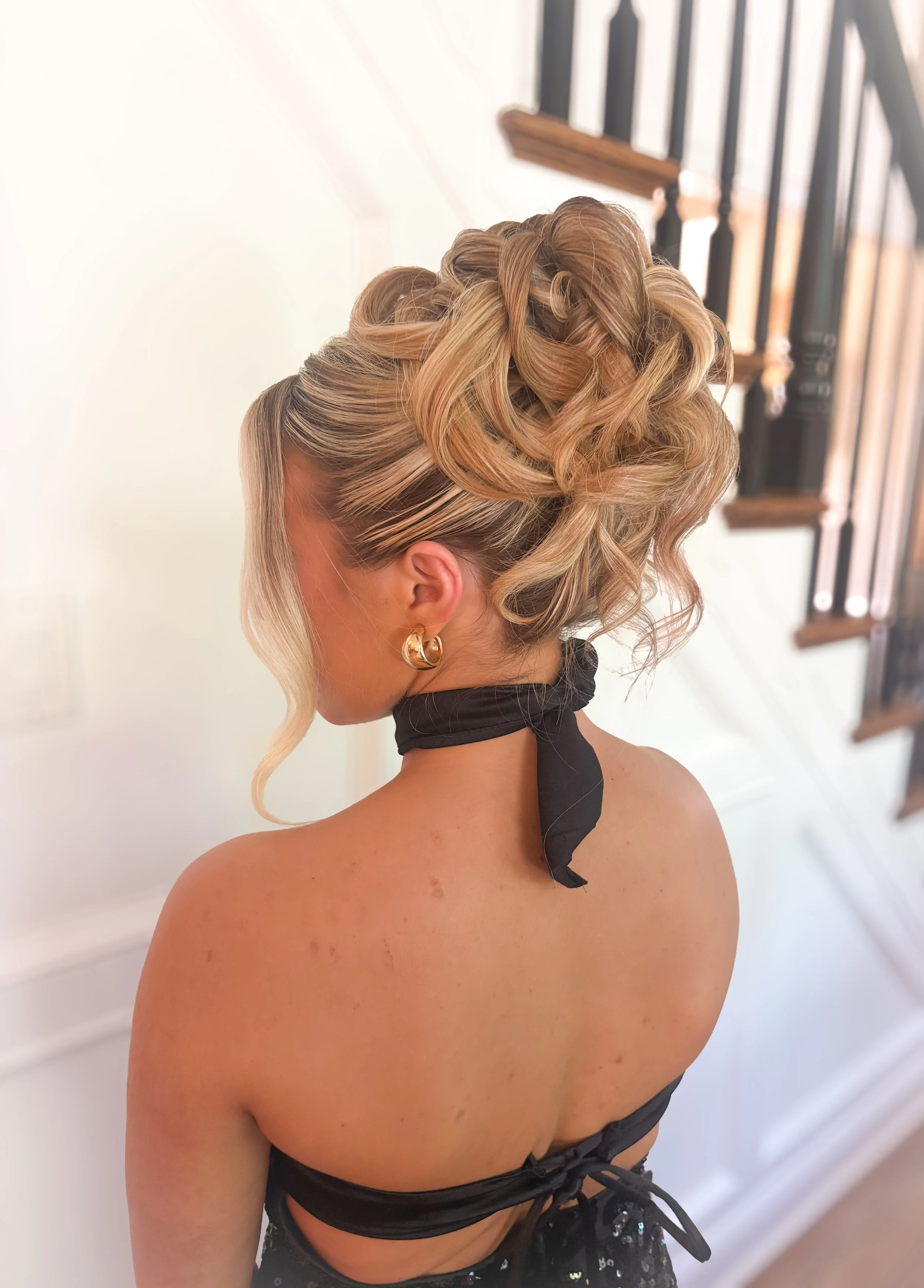 Woman with blonde curly hair styled in an elegant updo, wearing gold hoop earrings, a black choker, and a black strapless top or dress, standing near a staircase with wooden steps and black railing.