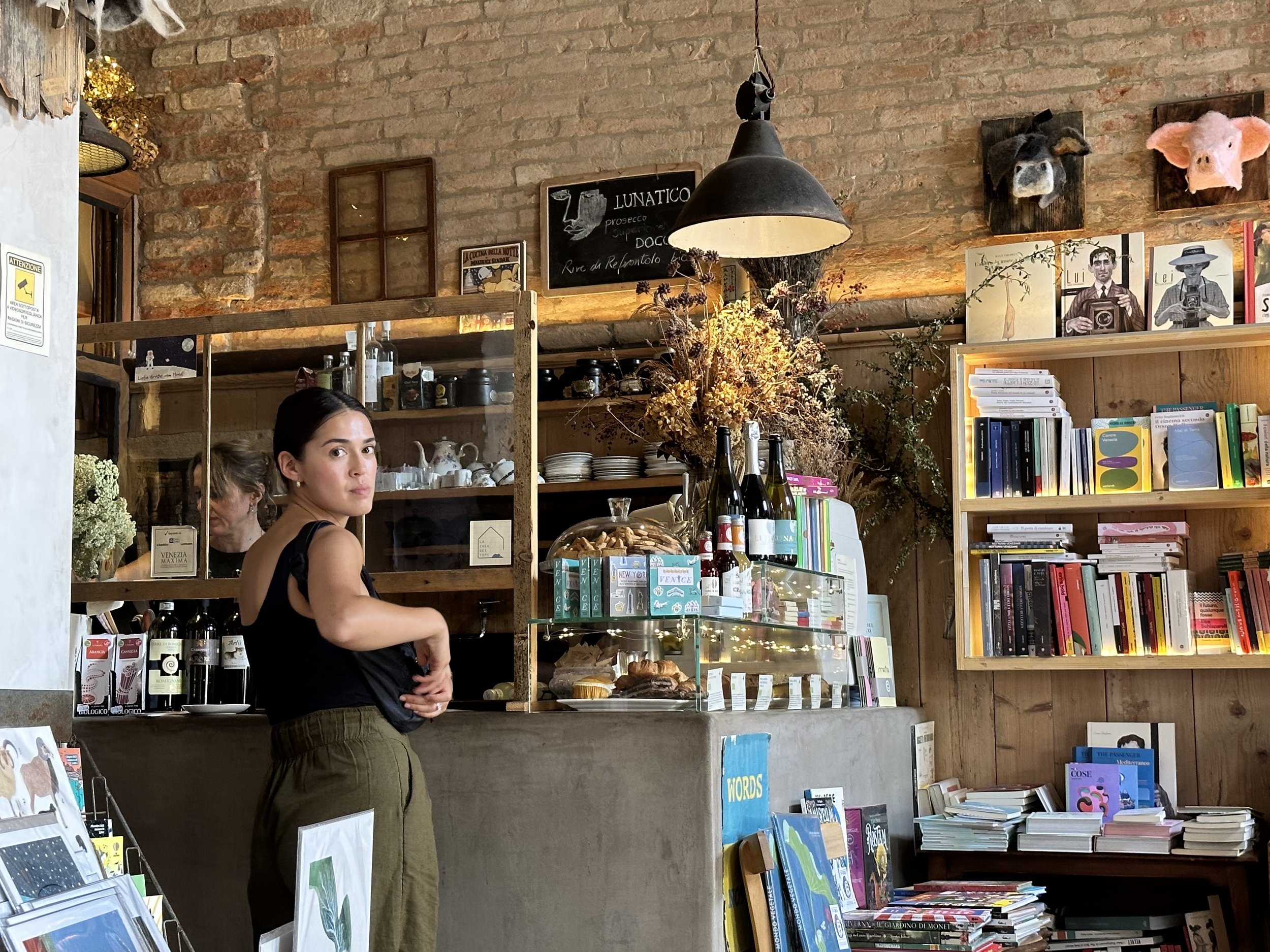 Interior of a bookstore or cafe with exposed brick walls, featuring a woman with dark hair, wearing a black top and green pants, standing near a display case with baked goods and magazines. Decor includes animal head wall mounts, artwork, a chalkboard menu, and bookshelves with colorful books.