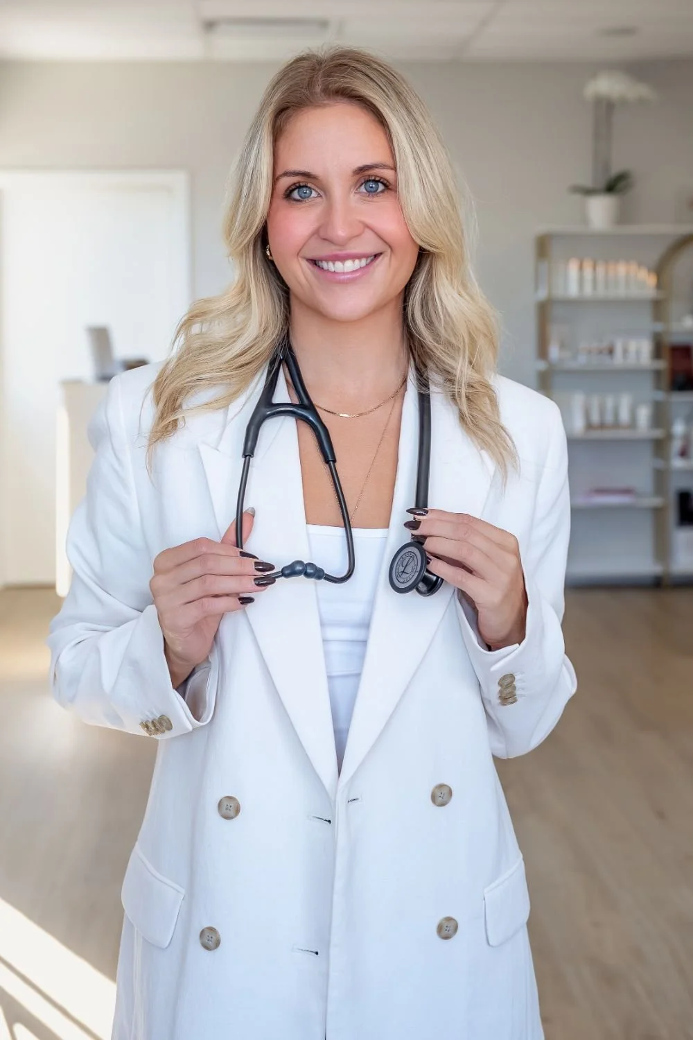 A smiling female doctor or healthcare professional with blonde hair, wearing a white coat and holding a stethoscope in a medical office or clinic.