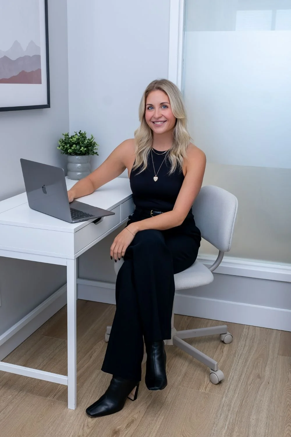 A woman with blonde hair in a black sleeveless top and black pants, sitting in an office chair at a white desk with a MacBook laptop. There is a small potted plant on the desk and a framed abstract artwork hanging on the wall behind her.