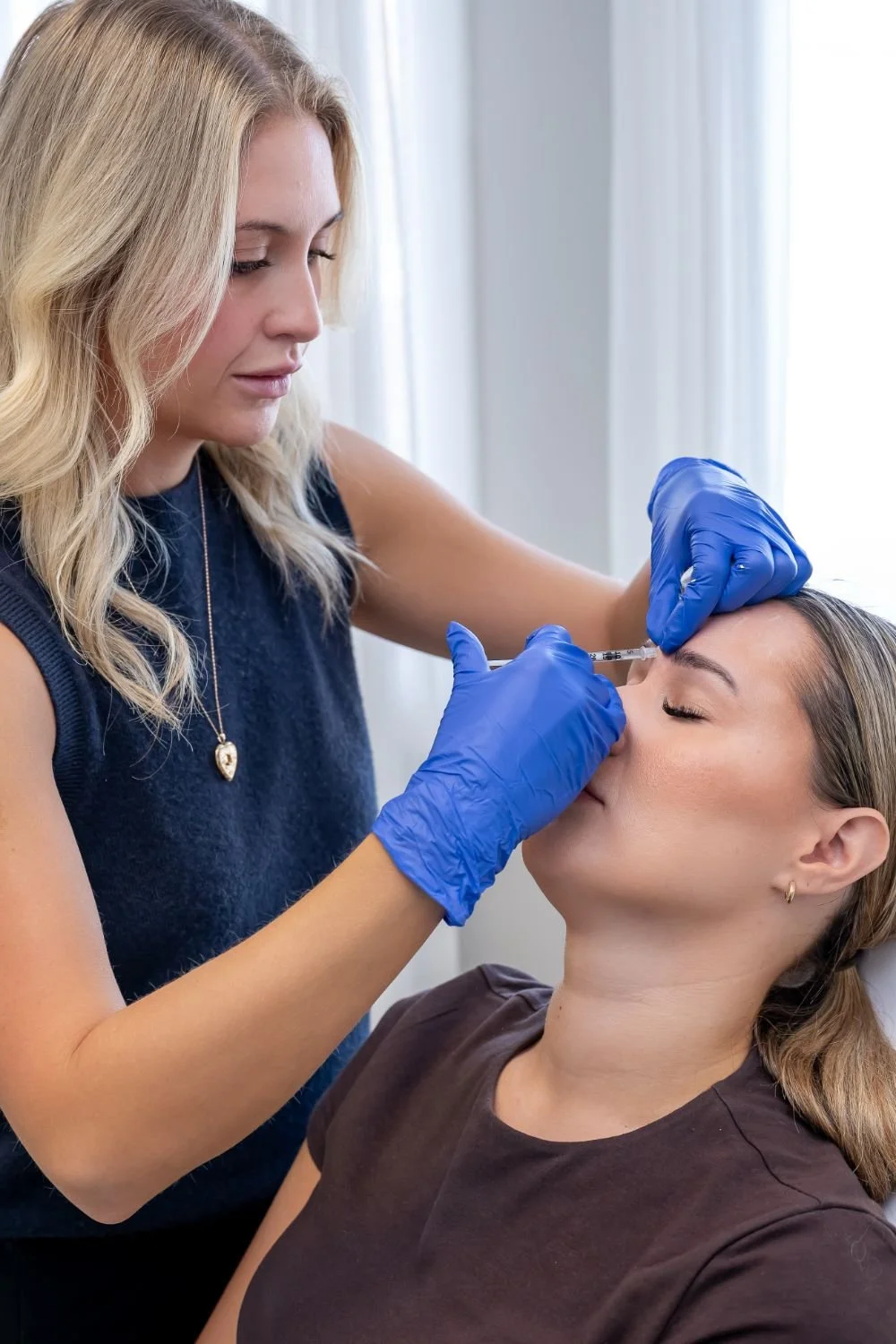 A woman lying down with her eyes closed receives a cosmetic injection in her forehead from a professional wearing blue gloves.