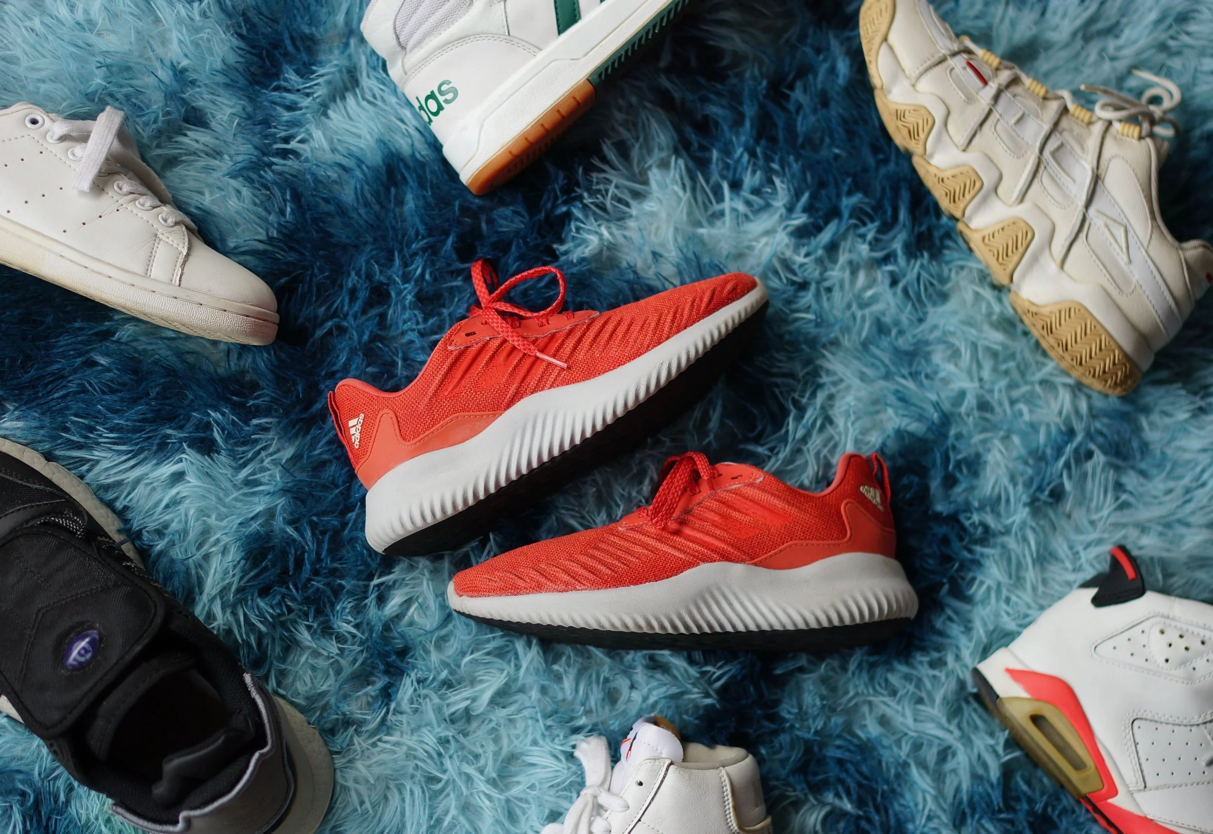 A sneaker collection laid out on a blue shag rug, with a pair of red-orange adidas running shoes in the center and white and black sneakers around them, ready to be cataloged before a move.