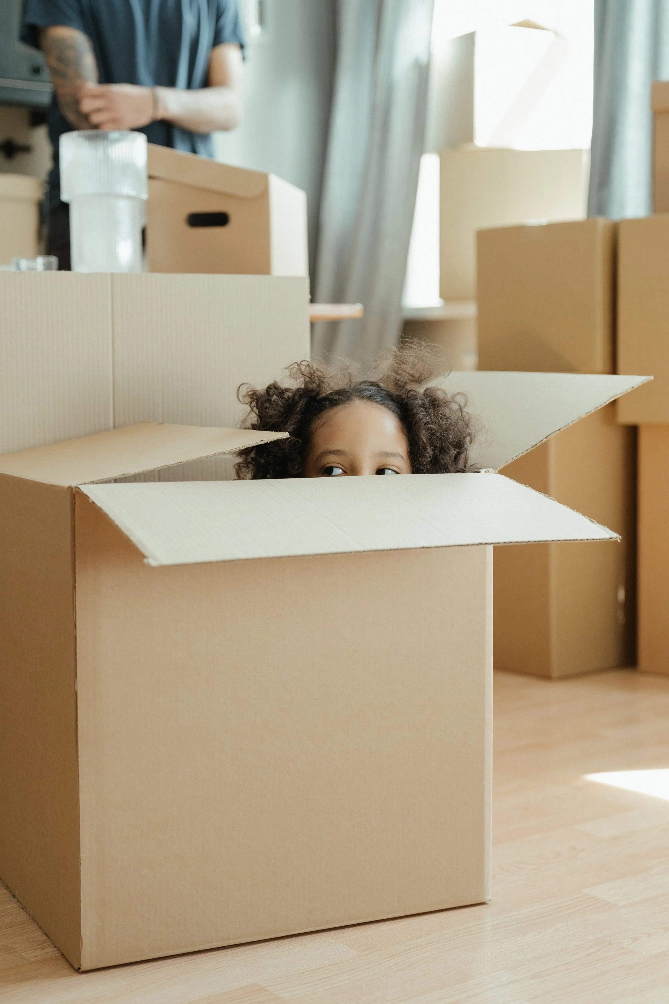 A young child peeking out from inside a large moving box while an adult packs in the background — capturing the playful reality of packing for a move with kids at home, with support from Home to Home Services in Richmond, VA
