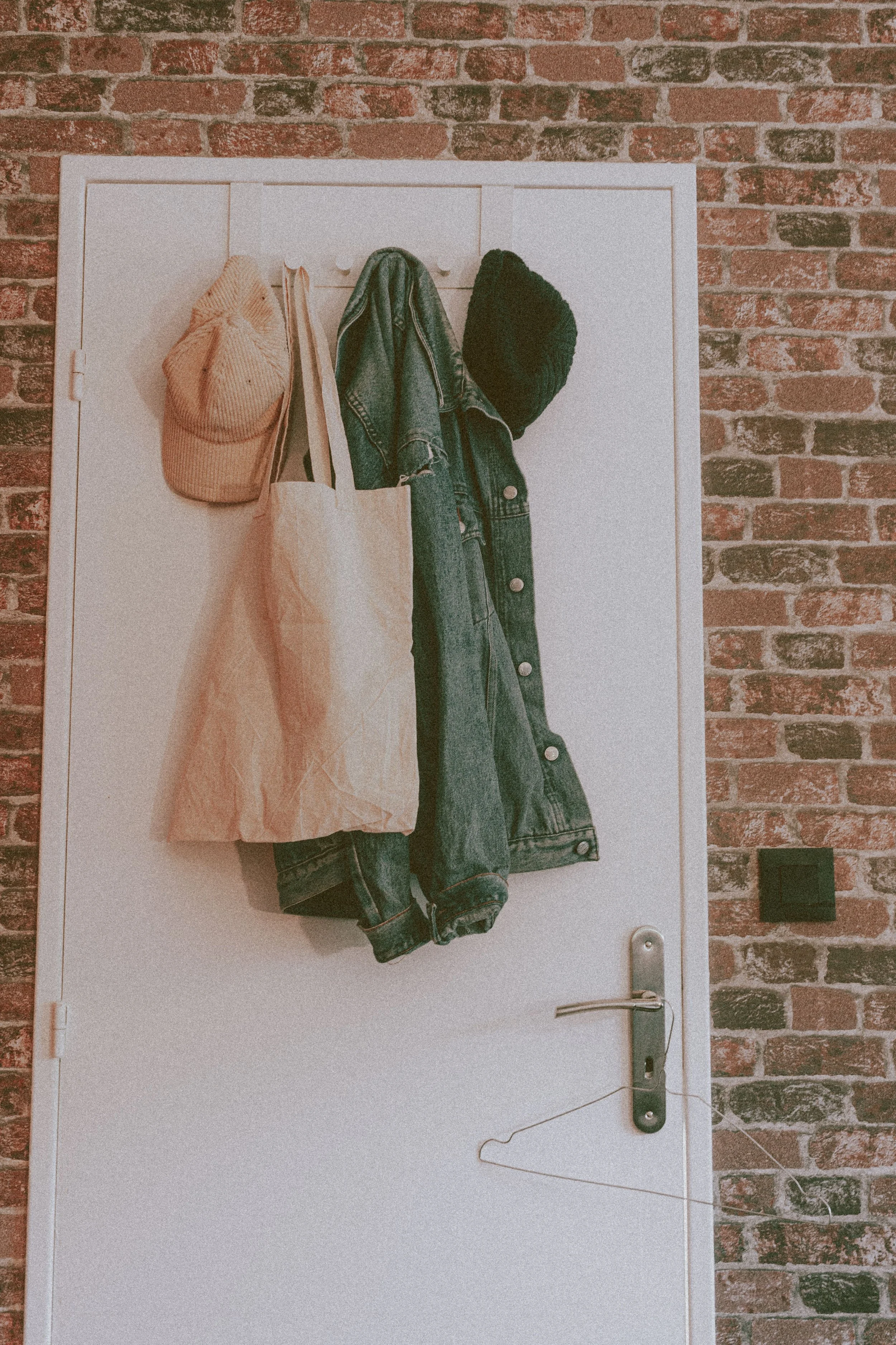 A white door with wall-mounted hooks holding a denim jacket, tote bag, baseball cap, and hat against an exposed brick wall — demonstrating door-mounted storage as a space-maximizing solution for small homes, from Home to Home Services in Richmond, VA