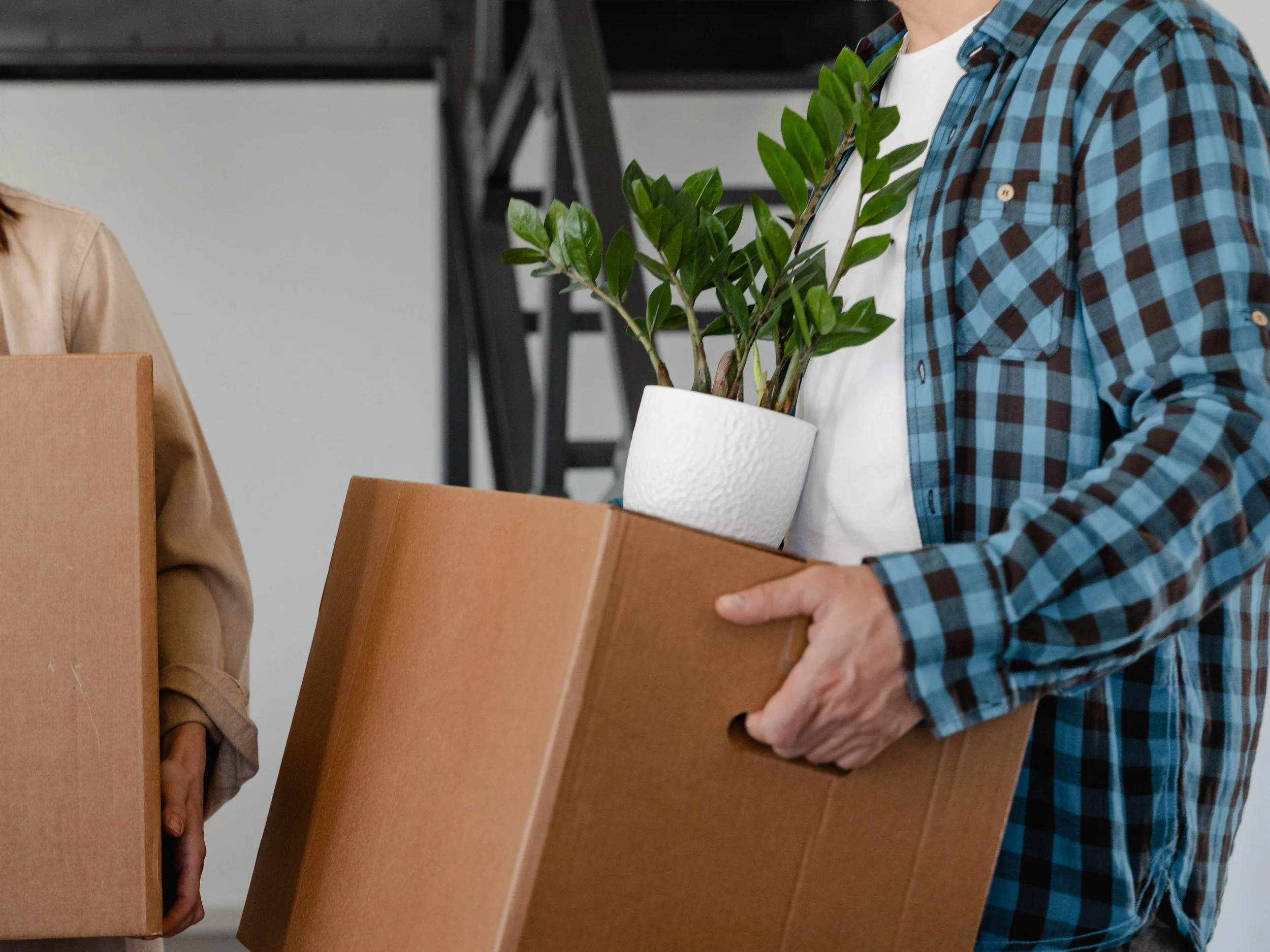 Two people carrying moving boxes — one holding a box with a potted plant tucked inside — representing the intentional, right-sizing approach to downsizing a home supported by Home to Home Services in Richmond, VA and Washington, DC