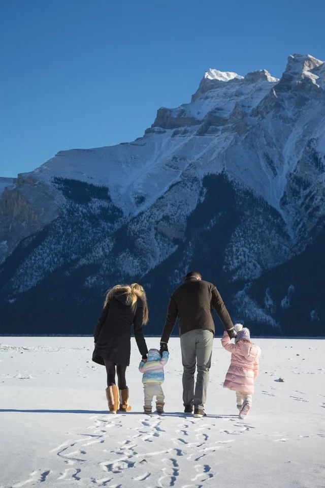 Family of four with two small children, all walking in the snow leaving footprints with mountains in the background