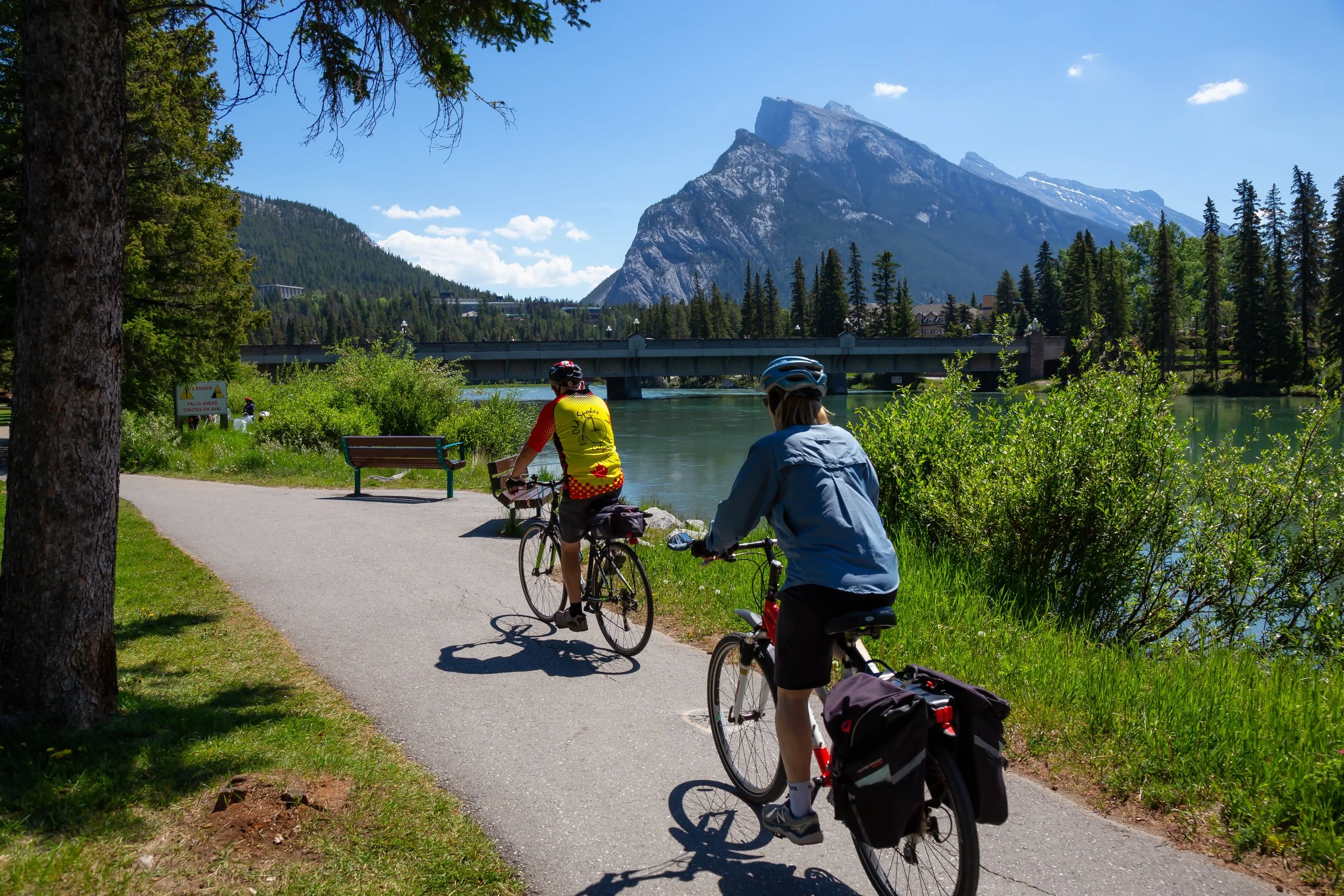 Two bike riders on a paved path next to a river, mountain in the background