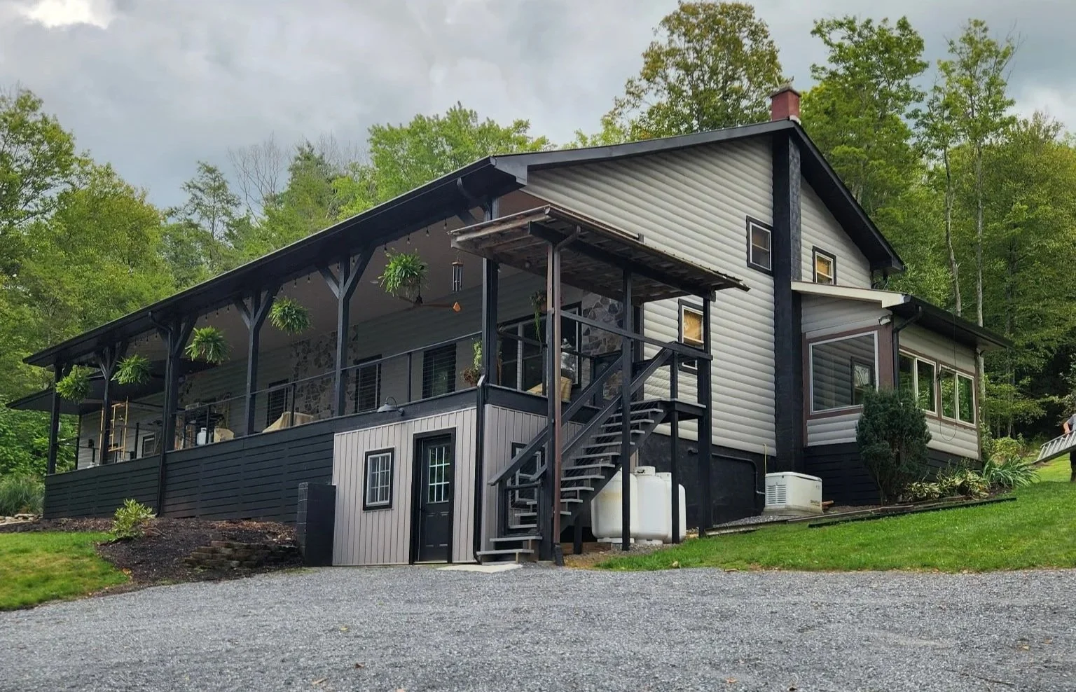 Modern two-story house with a large deck, hanging plants, and a gravel driveway, surrounded by green trees under a cloudy sky.