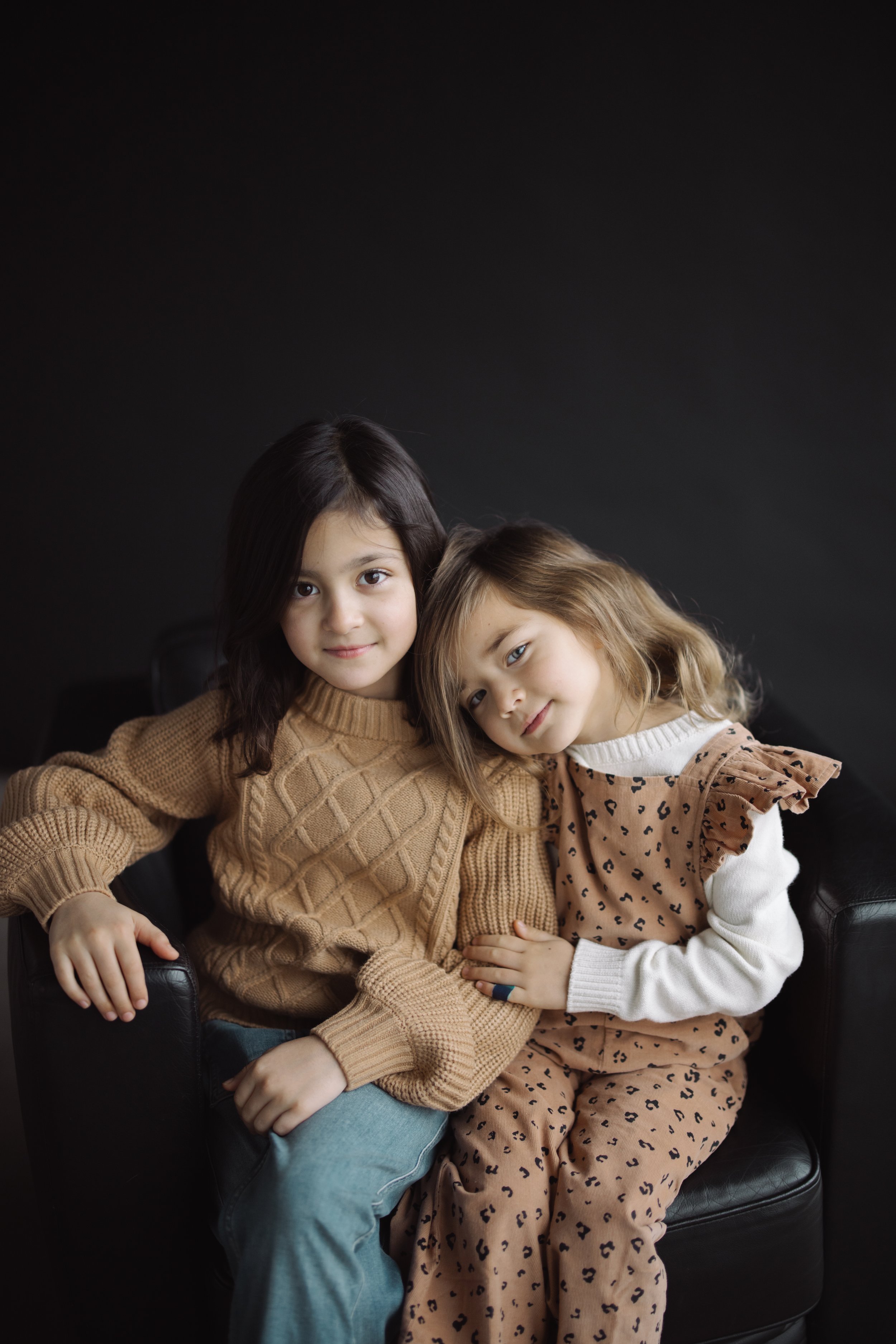 Two young children sitting closely on a black chair, one wearing a brown knit sweater and the other in a patterned dress, against a dark background.