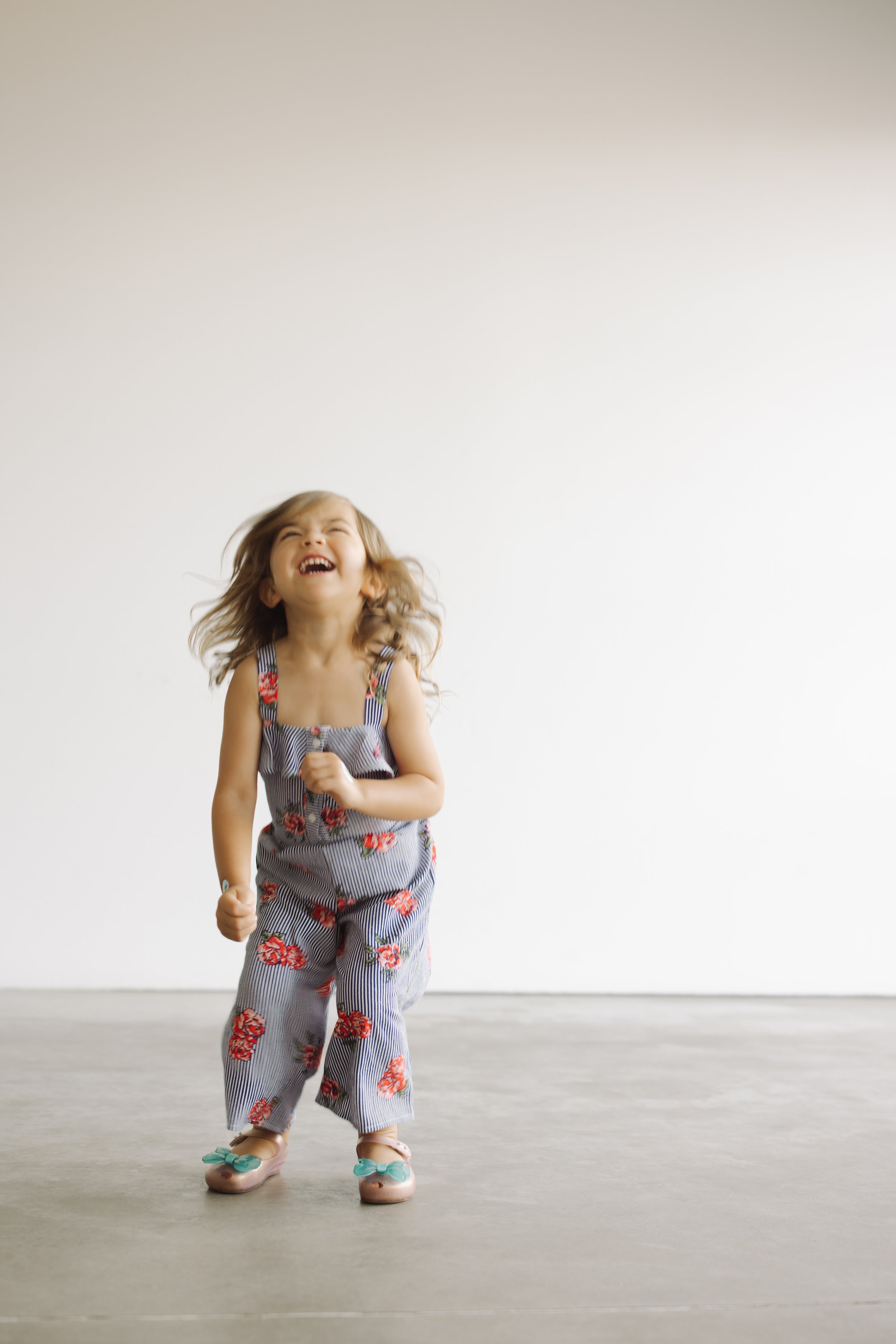 Young child in floral overalls laughing while jumping indoors, wearing pink shoes with bows.