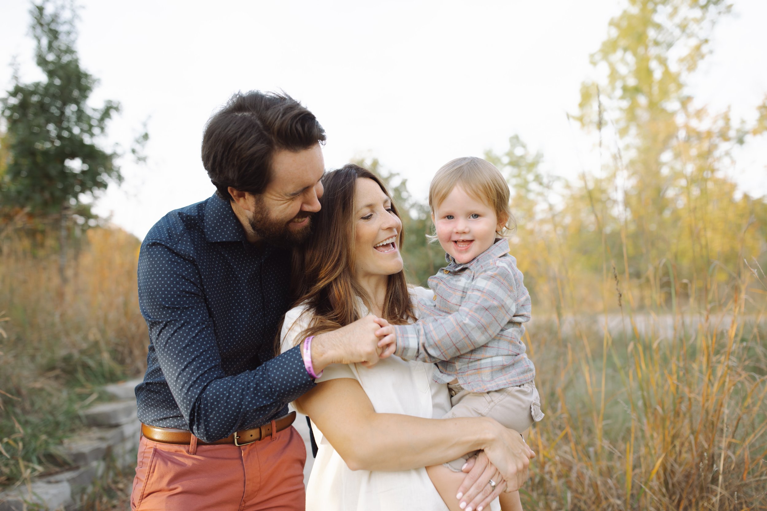 A family of three smiling in a field, with tall grass and trees in the background. The father and mother are holding a toddler.