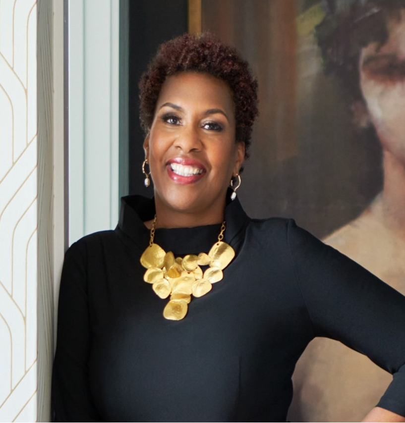 A smiling Black woman with short curly hair, wearing a black top, gold statement necklace, and matching earrings, standing indoors next to a window and a dark background with artwork.