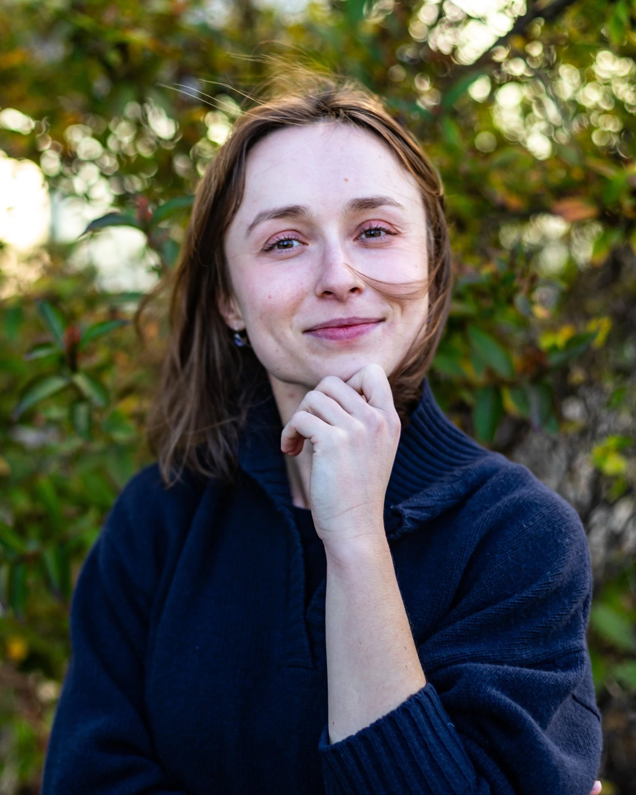A young woman with light skin and brown hair outside, smiling and resting her chin on her hand, wearing a dark blue hoodie.
