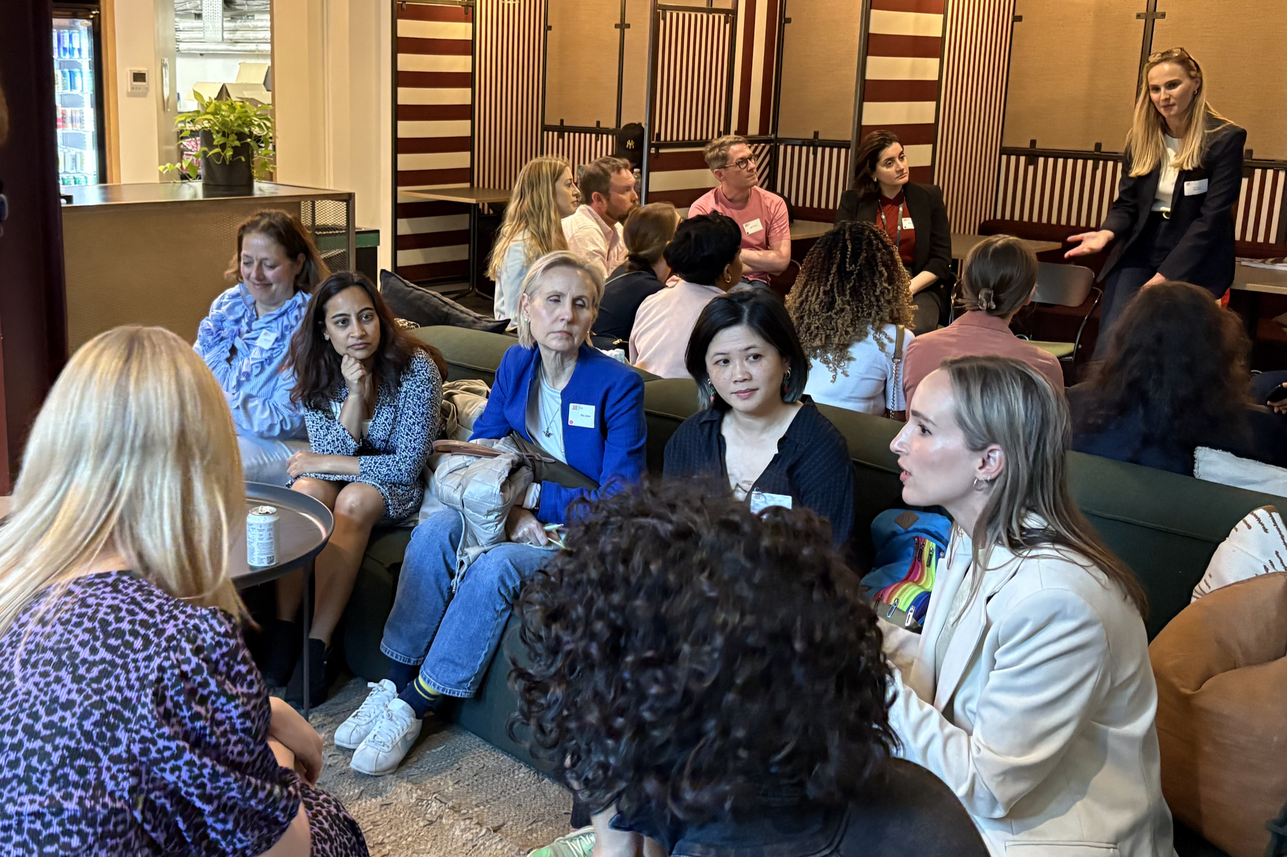 A woman standing and speaking to a group of seated women in a lounge or conference room setting.