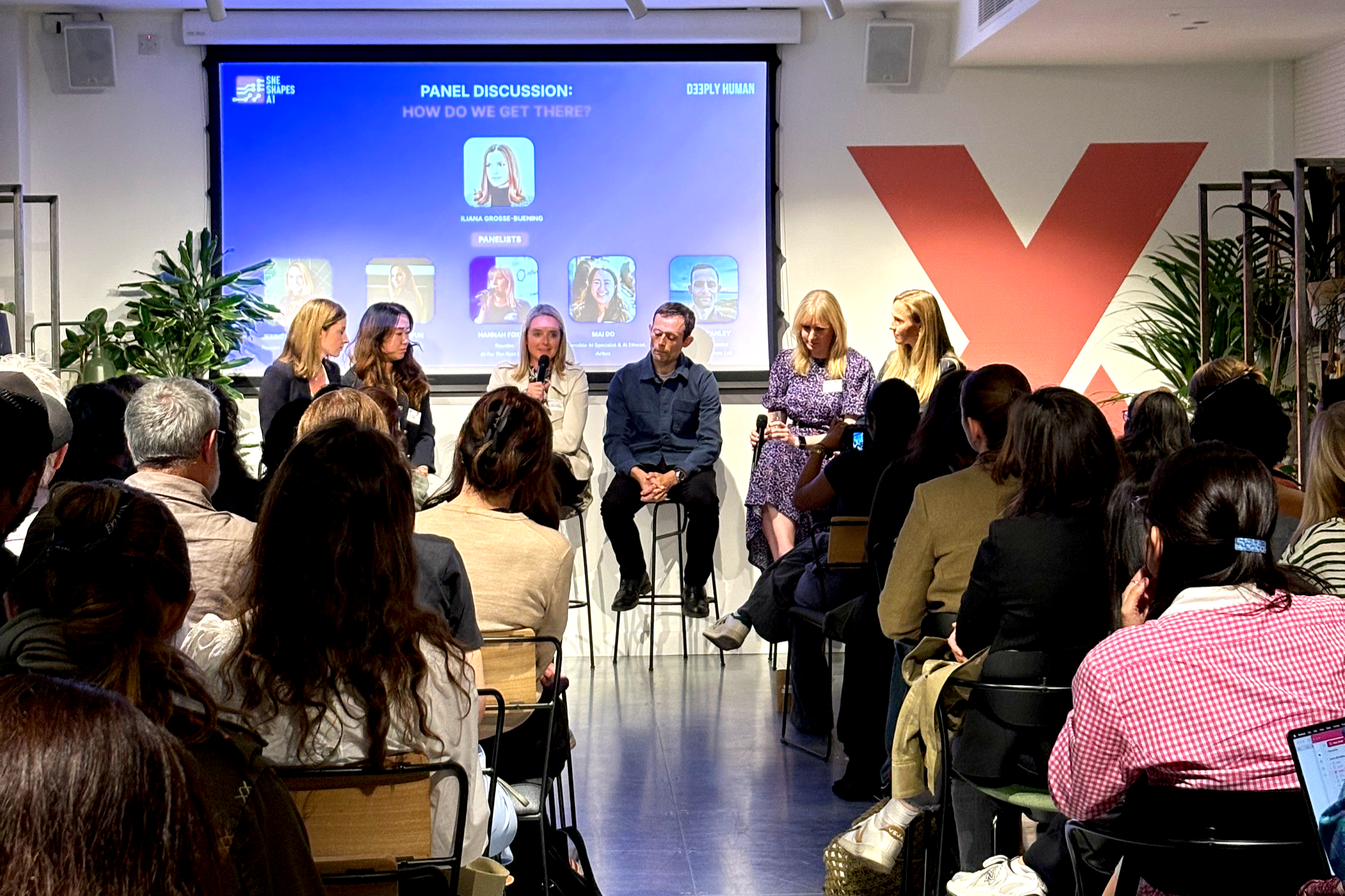 A panel discussion with seven women and one man sitting on stage in front of an audience at a conference or event. A large screen behind them displays the panel's details and names.