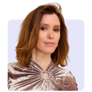 A woman with shoulder-length light brown hair, wearing a silver-colored top with a ring detail at the neckline, standing against a light background.