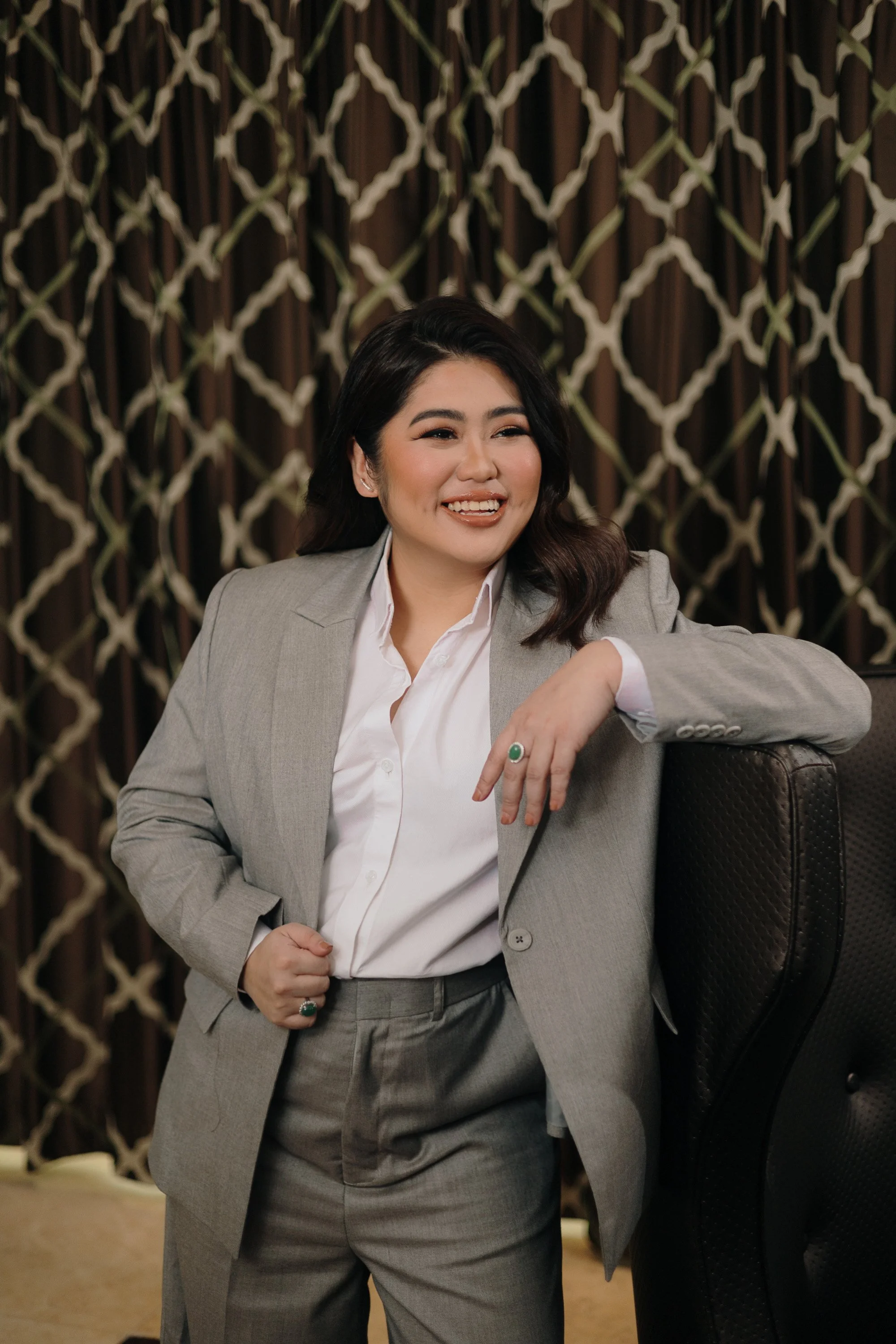 A woman in a grey suit smiling and leaning against a black chair, with a patterned brown background.
