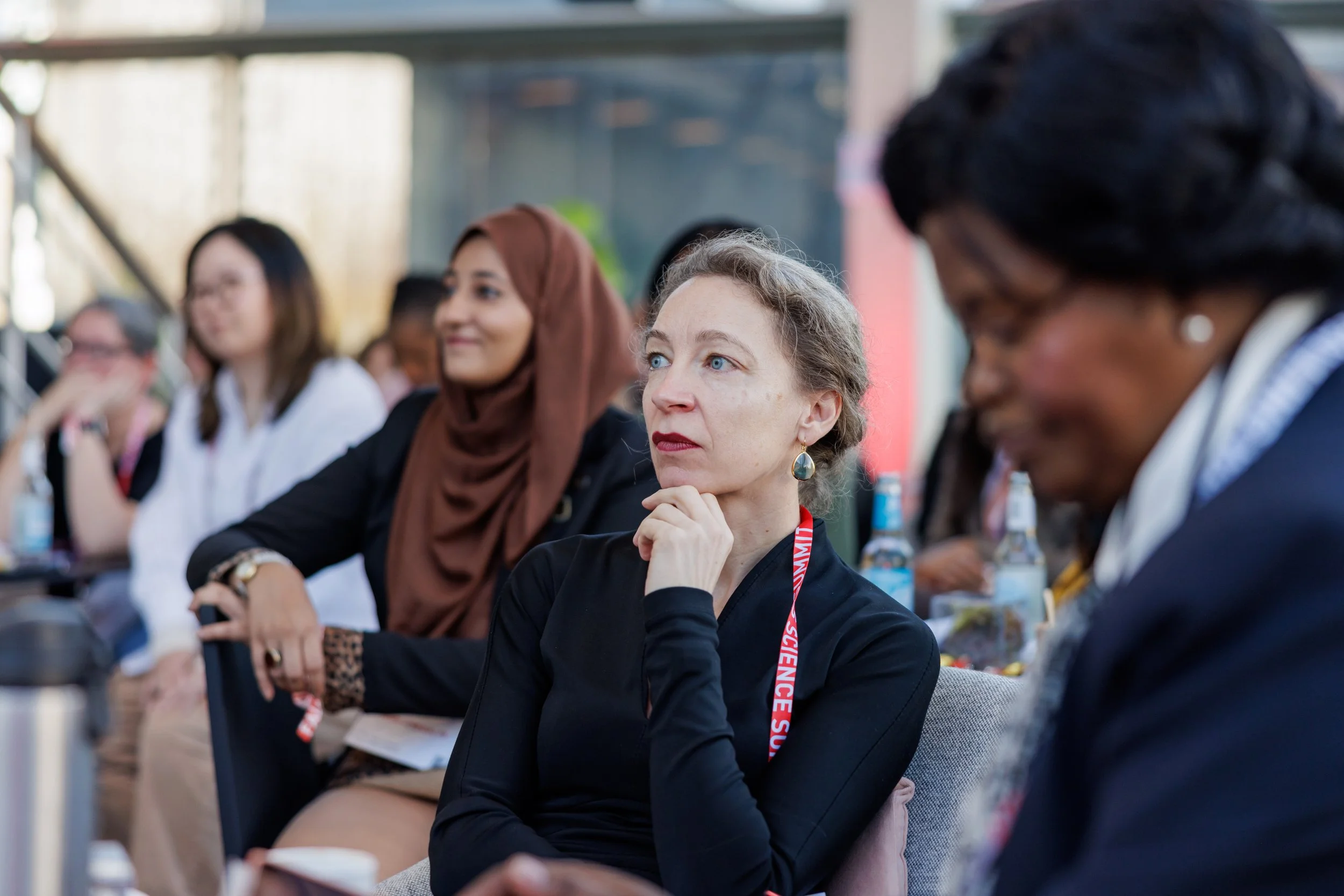 A group of diverse women attending a conference, sitting attentively, with one woman in focus resting her chin on her hand, wearing a black top and a red conference badge.