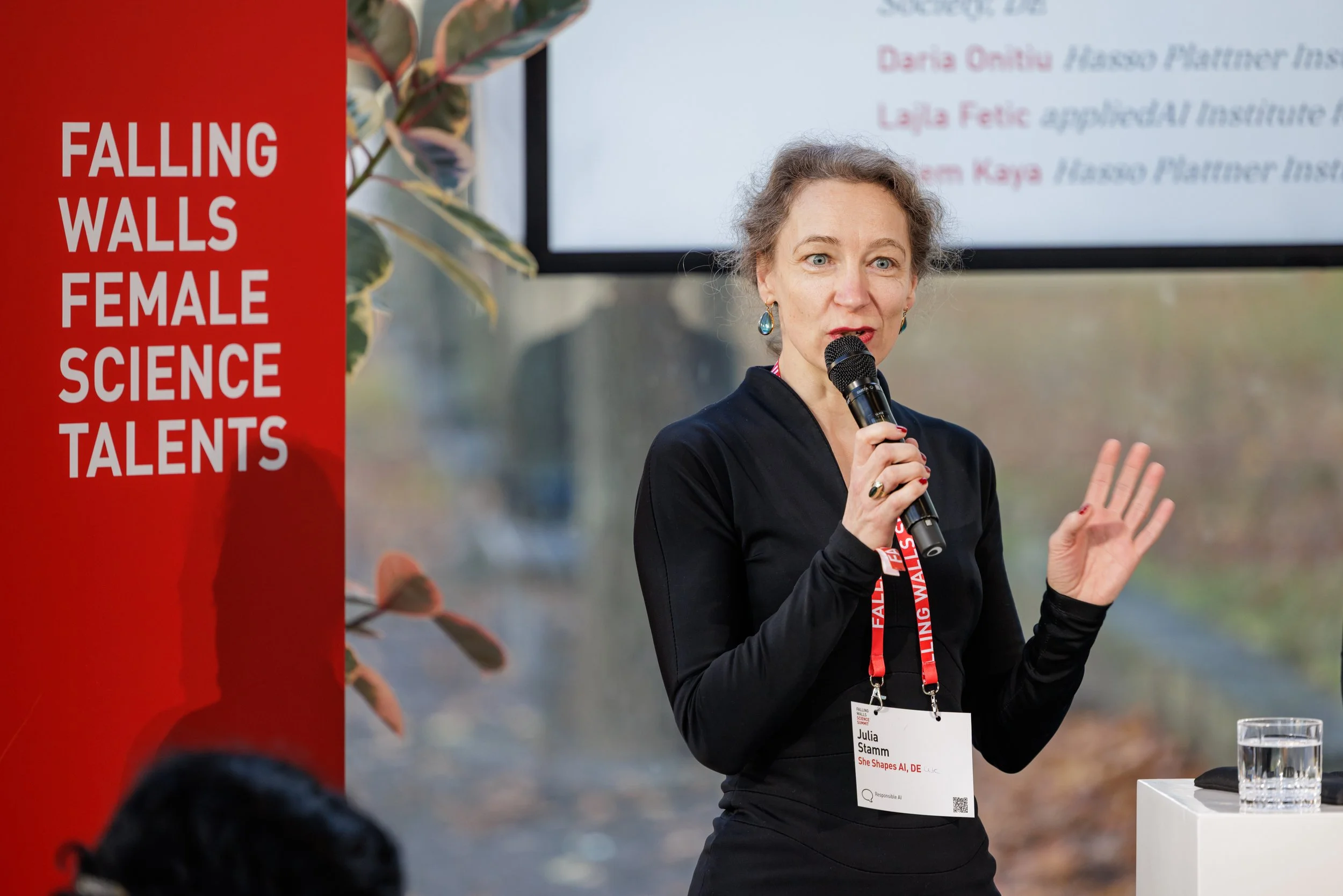A woman speaking into a microphone at a conference, with a banner reading 'Falling Walls Female Science Talents' and a large screen behind her displaying her name, Julia Stamm, and her affiliation, She Shapes AI, DE.