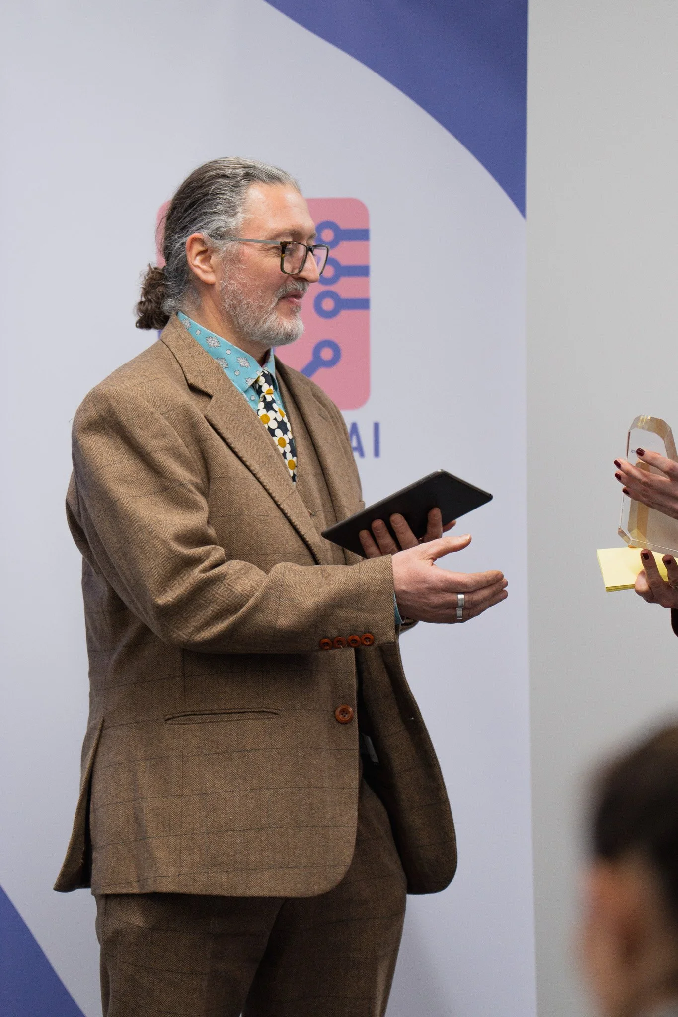 A man with gray hair, glasses, and a beard, dressed in a brown suit, is holding a tablet while talking to someone at a professional event or conference.