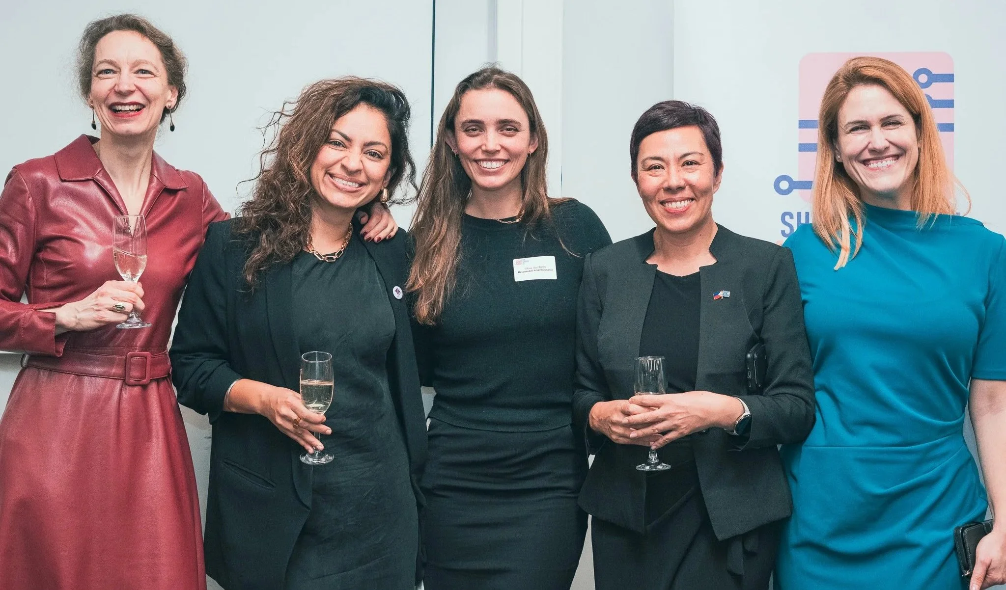 Six women smiling and holding glasses of champagne at a professional event, standing in front of a wall with a QR code logo.