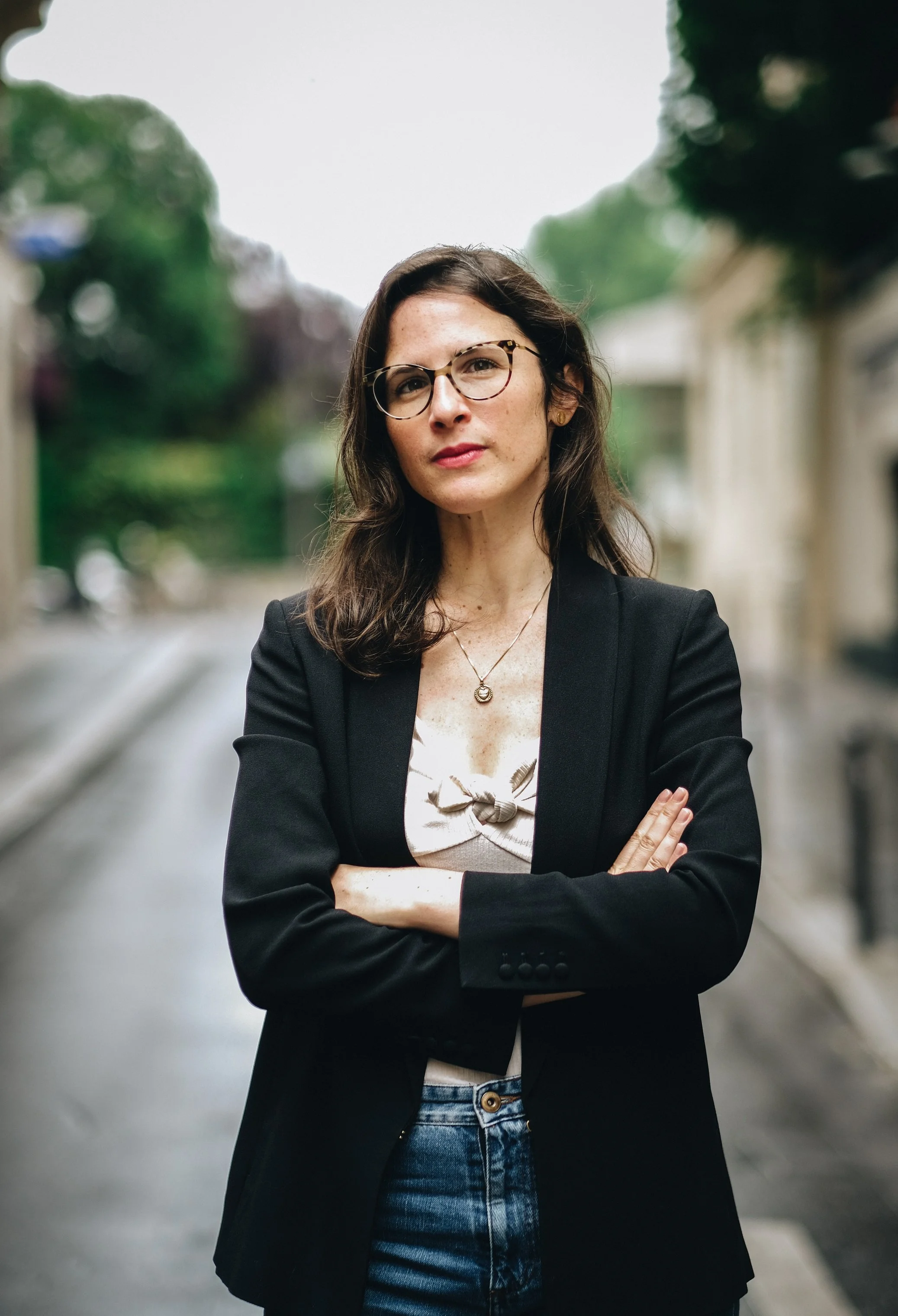 A woman with brown hair, glasses, and a serious expression, crossing her arms on a city street during the day.