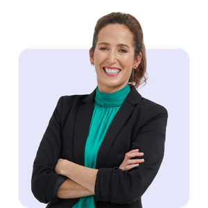 A woman smiling with arms crossed, wearing a black blazer and a teal blouse, standing against a light-colored background.