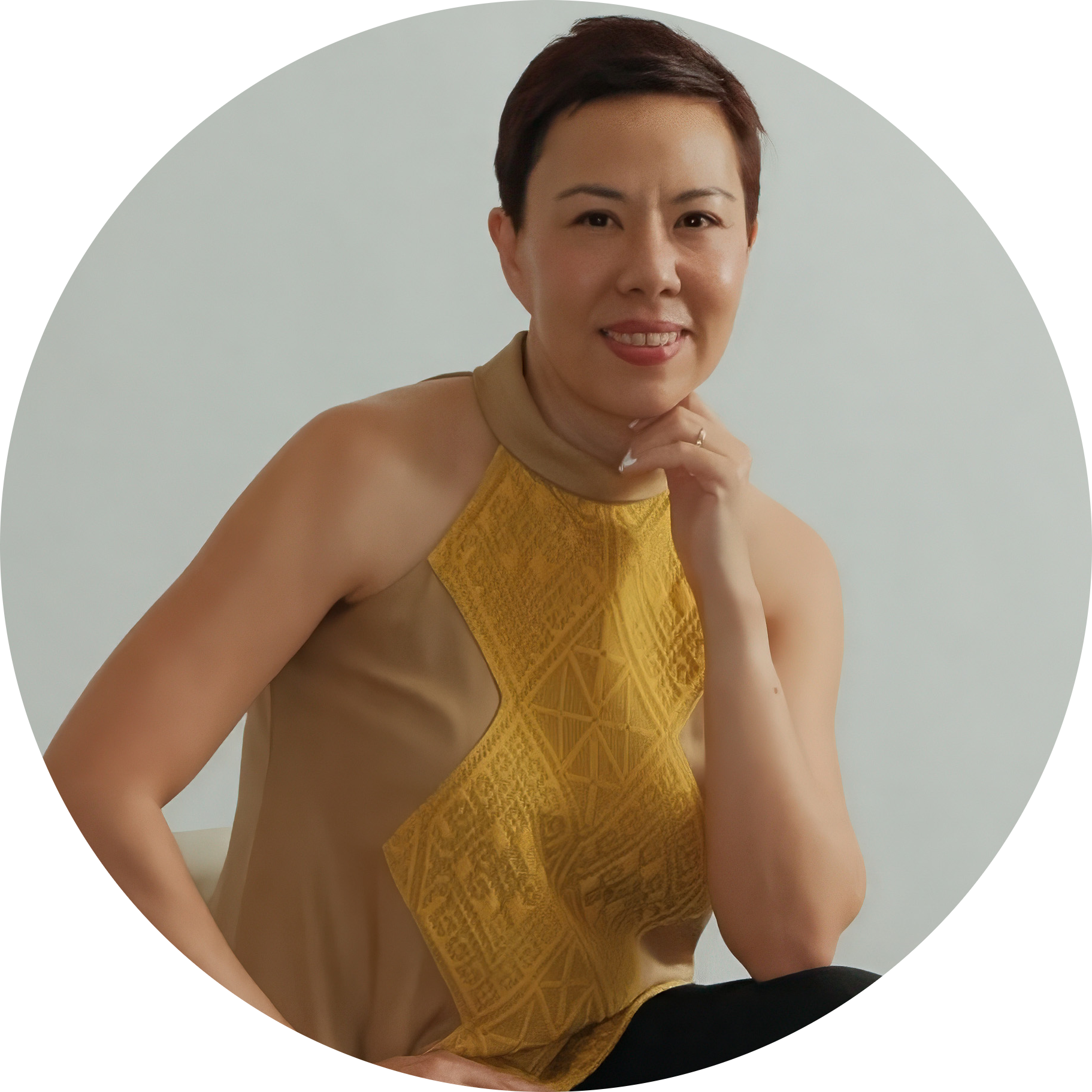 A woman with short dark hair, wearing a beige and yellow sleeveless top, sitting in front of a light-colored wall, smiling and resting her chin on her hand.