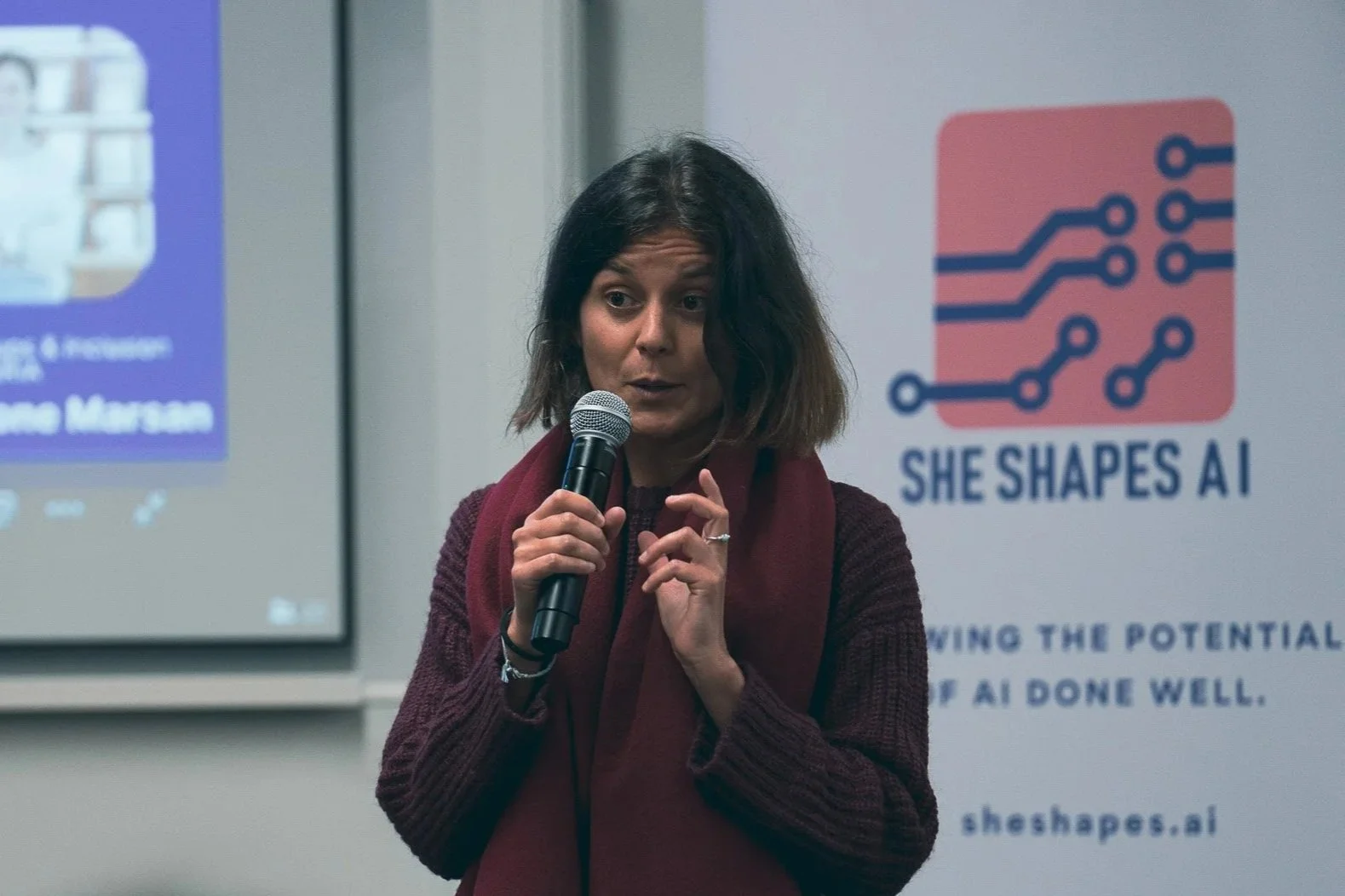 A woman speaking into a microphone at a presentation with a 'She Shapes AI' banner in the background.
