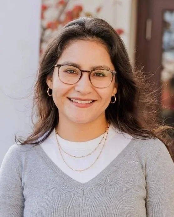 A young woman with long brown hair and glasses, smiling outdoors, wearing a light gray sweater and layered necklaces.