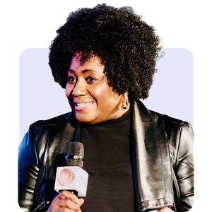 A woman with curly black hair holding a microphone, wearing a black leather jacket and black top, smiling against a light background.