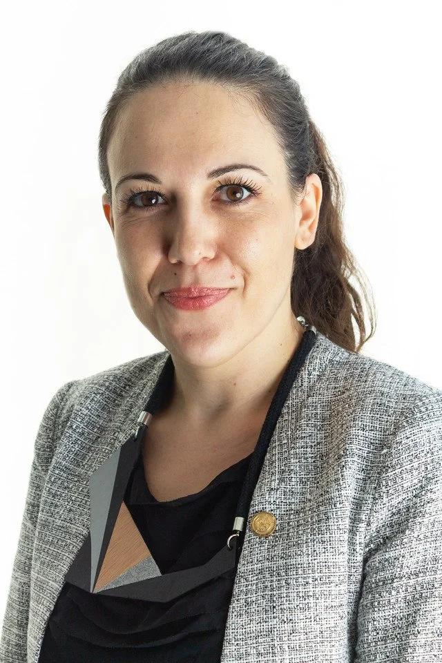 A woman with brown hair tied back, wearing a gray blazer and a black top, looking at the camera with a slight smile, standing against a white background.