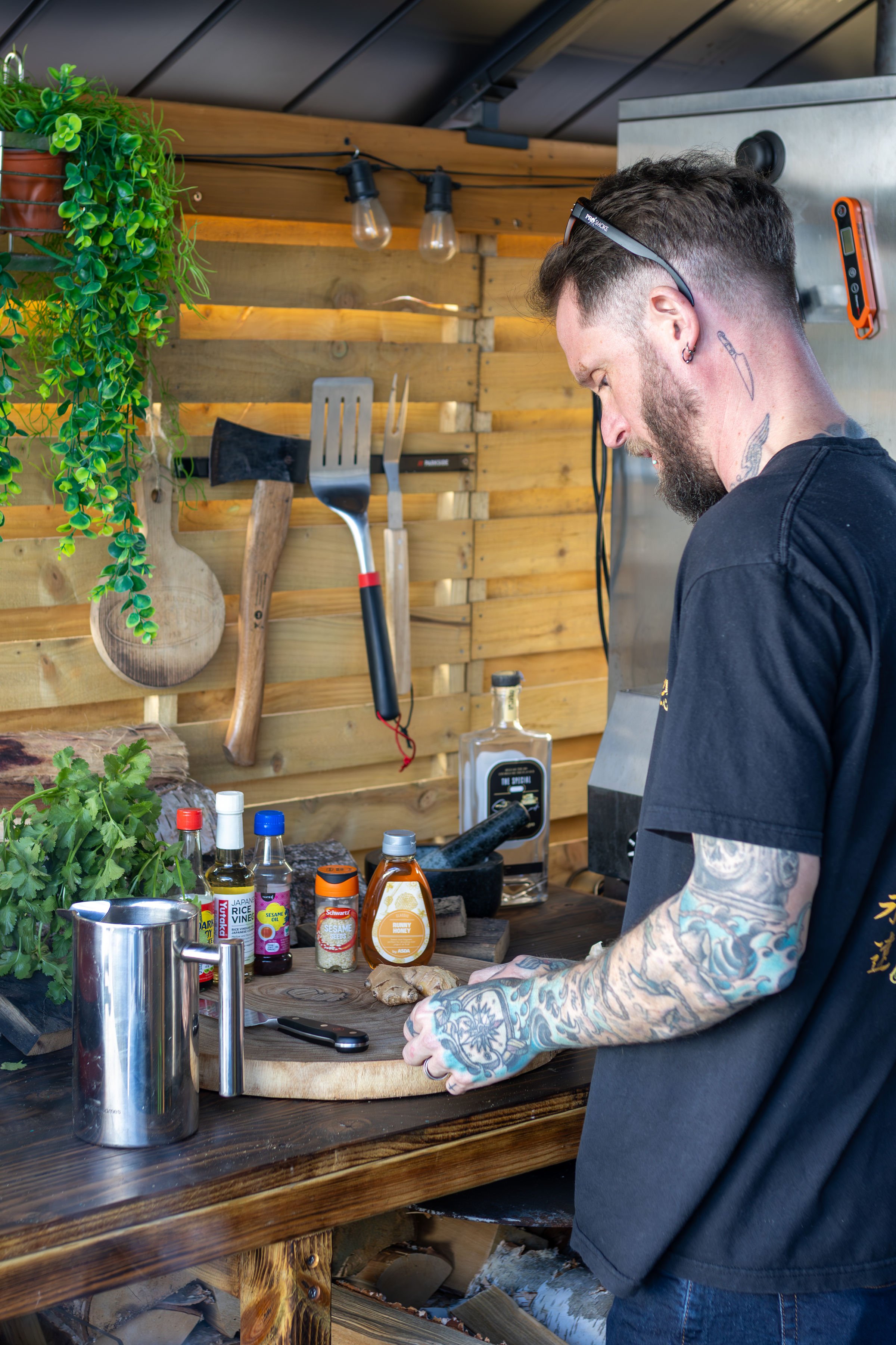 Man with tattoos cooking in a rustic outdoor kitchen with wooden walls, herbs, condiments, and kitchen tools.
