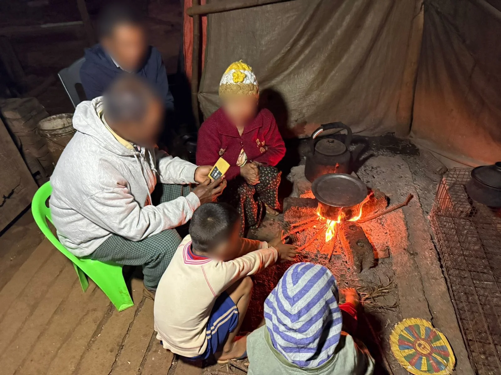 Family sitting around cooking fire inside tarp shelter listening to Treasure audio Bible