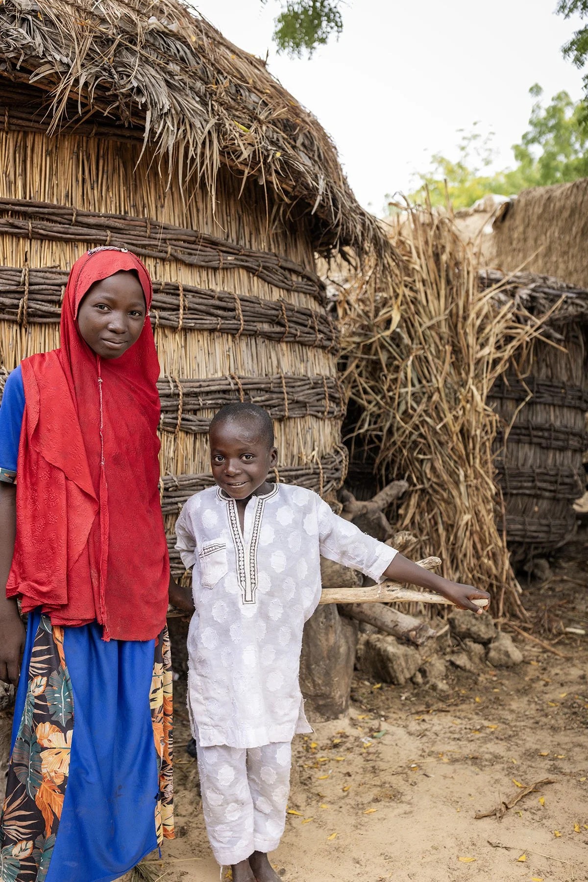 A young woman and a boy stand outdoors in front of a thatched hut. The woman wears a red headscarf and a red dress with blue sleeves, and the boy wears a white traditional outfit. They are smiling and the boy is pointing towards the ground with a stick.